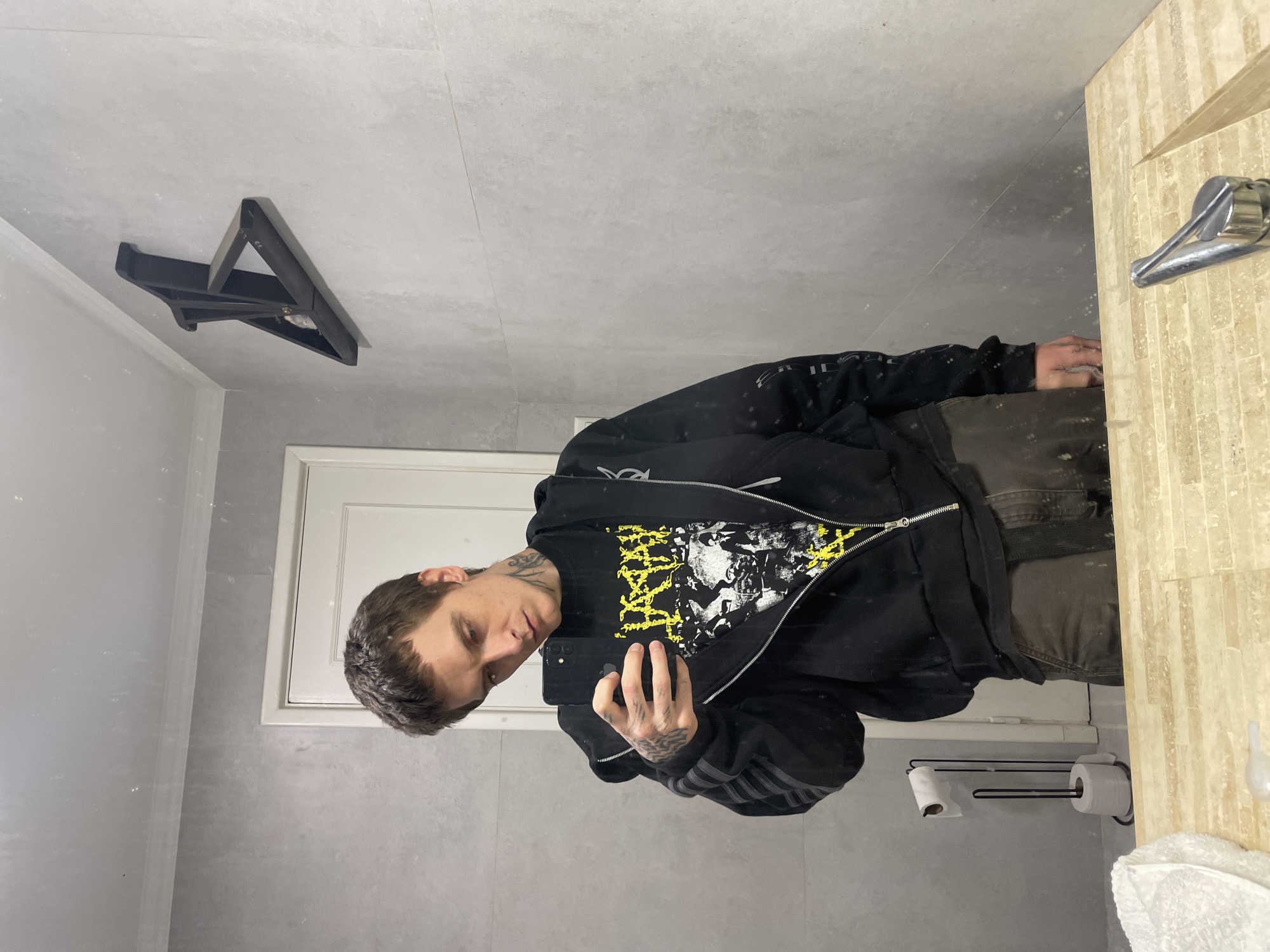 A person is taking a selfie in a bathroom with gray tile walls. The person is wearing a black zip-up hoodie with a graphic t-shirt underneath and dark-colored pants. The bathroom includes a mirror, a white door with a white frame, a black ceiling-mounted light fixture, a toilet paper holder, and a sink with tan striped tiles. The location is in Vicente López, Argentina.