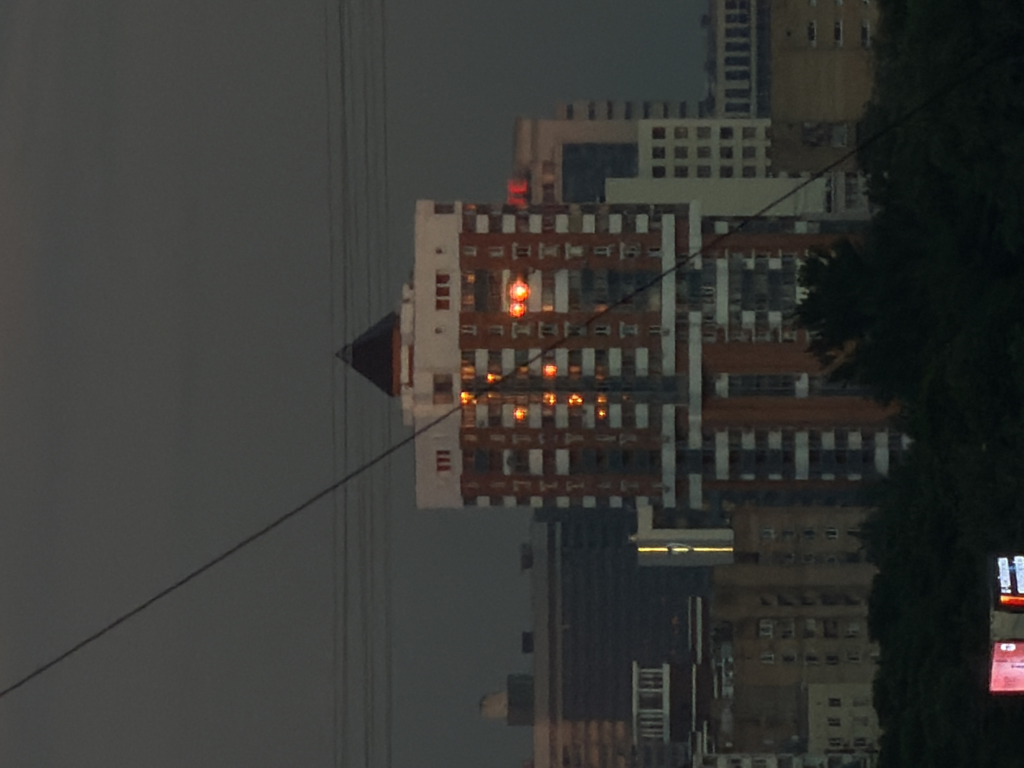 An exterior medium shot captures a cluster of buildings under a dark, overcast sky. The main subject is a tall, multi-story building with a distinctive red and white facade, adorned with orange lights on several floors. This building is topped with a dark, pyramidal roof. To its left and right are other buildings of varying heights and architectural styles, some with visible windows and balconies. A diagonal dark line, possibly a wire or cable, cuts across the upper left portion of the frame. In the foreground, a dense line of dark green trees partially obscures the lower portion of the buildings. At the very bottom of the frame, a small section of a brightly lit red sign is visible. The overall impression is an urban scene at dusk or twilight.