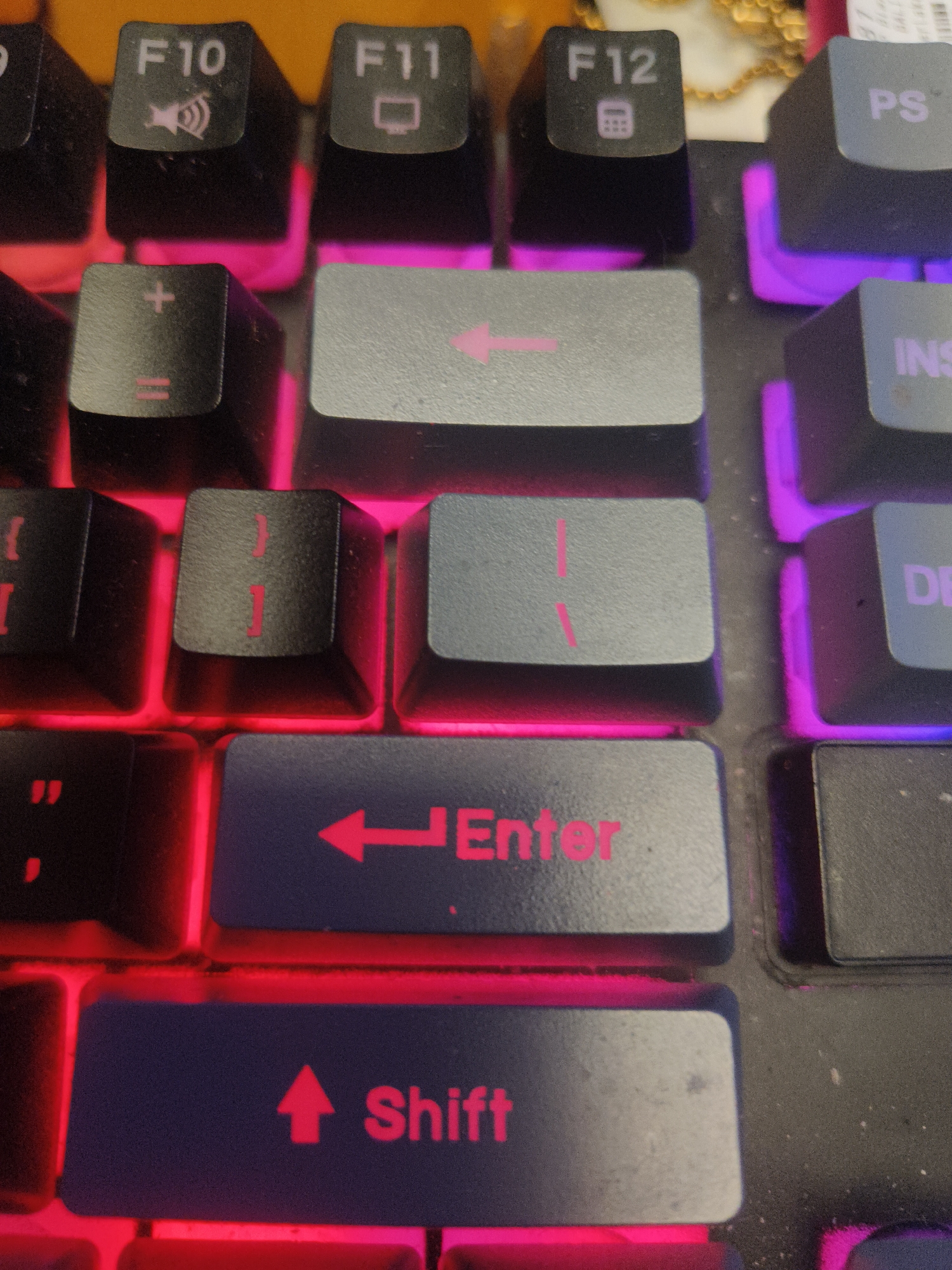 The image shows a close-up, eye-level view of a section of a black gaming keyboard with RGB backlighting, illuminated in red and purple. The keys are made of dark plastic with white lettering and symbols. Visible keys include "F10" with a speaker icon, "F11" with a monitor icon, "F12" with a calculator icon, "PS," "INS," and "DEL." Below these are keys with symbols like "+," "=", left arrow, and the word "Enter" with a left arrow. The bottom visible key shows the word "Shift" with an upward arrow. The lighting suggests a low-light environment, typical of gaming setups. There are no people or other objects visible in the frame. The image does not contain any information about the location in the city of Kuta Selatan, Indonesia, or any other geographical information.