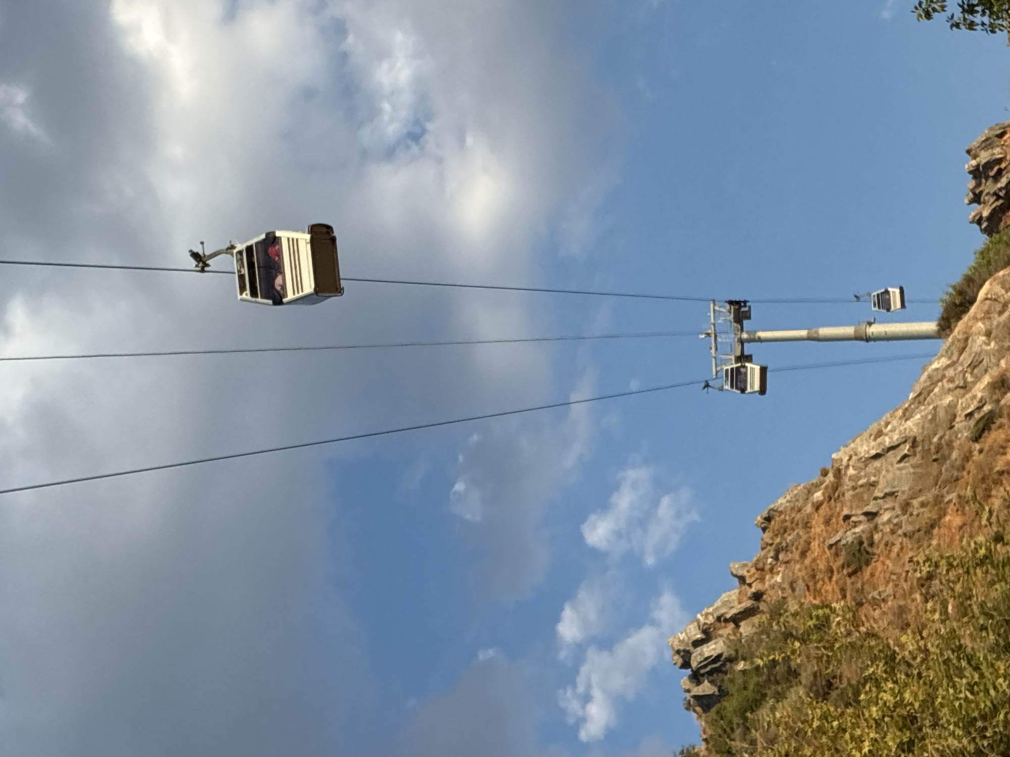 This image captures a scenic view of the Alanya Cable Car system in Alanya, Turkey, with its distinctive white cabins suspended against a bright blue sky dotted with white clouds. The cable car, a popular tourist attraction, ascends the side of a rugged, rocky mountain that occupies the left portion of the frame. The mountain exhibits a mix of rocky outcrops, sparse vegetation, and areas of dry, reddish-brown earth, indicative of a Mediterranean climate.
In the foreground, the jagged edges of the mountain are clearly visible, with some greenery clinging to its slopes. The cable car system consists of multiple cabins and supporting pylons. The main subject is a white cable car cabin with tinted windows, positioned mid-air and showing several passengers inside, including one person with red hair. It is suspended from a thick, dark cable. Further down the cable, another, smaller cable car is visible, also suspended in the air, and below that, a third, even smaller cabin can be seen. One of the pylons supporting the cable system is visible, a tall, gray structure with a white top.
The sky is a vibrant blue, with fluffy white cumulus clouds scattered across it, some casting soft shadows. The lighting suggests it is daytime, likely late morning or early afternoon, given the brightness and the clear visibility of the scene. The overall impression is one of a functional piece of infrastructure integrated into a natural, mountainous landscape, offering visitors panoramic views of Alanya and the surrounding coastline.