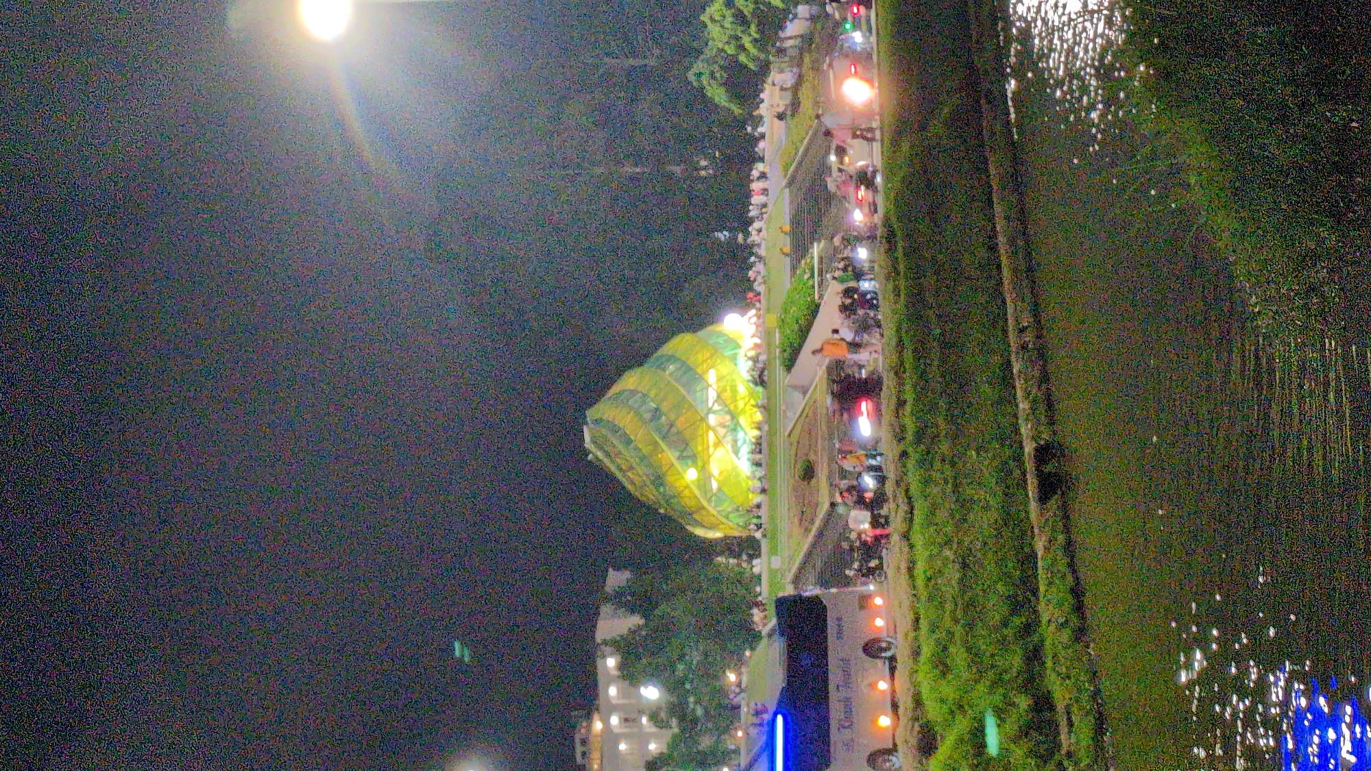 This is a nighttime, slightly grainy, vertical, medium-distance, full-color photograph of the Da Lat Flower Garden in Da Lat, Vietnam. In the center of the frame is the iconic Da Lat Pedagogical College building, a distinctive conical structure with green and yellow geometric patterns that illuminate the night. A crowd of people, some on motorbikes, are gathered around the building and on the steps leading up to it. In the foreground, a white bus with blue lights on top is parked to the left, with the text "Khanh Tourist" visible on its side. A grassy embankment runs along the bottom of the image, with a body of water reflecting lights from the scene. Trees and other buildings are visible in the background, silhouetted against the dark night sky. A bright streetlamp shines from the upper right corner, creating a lens flare effect.