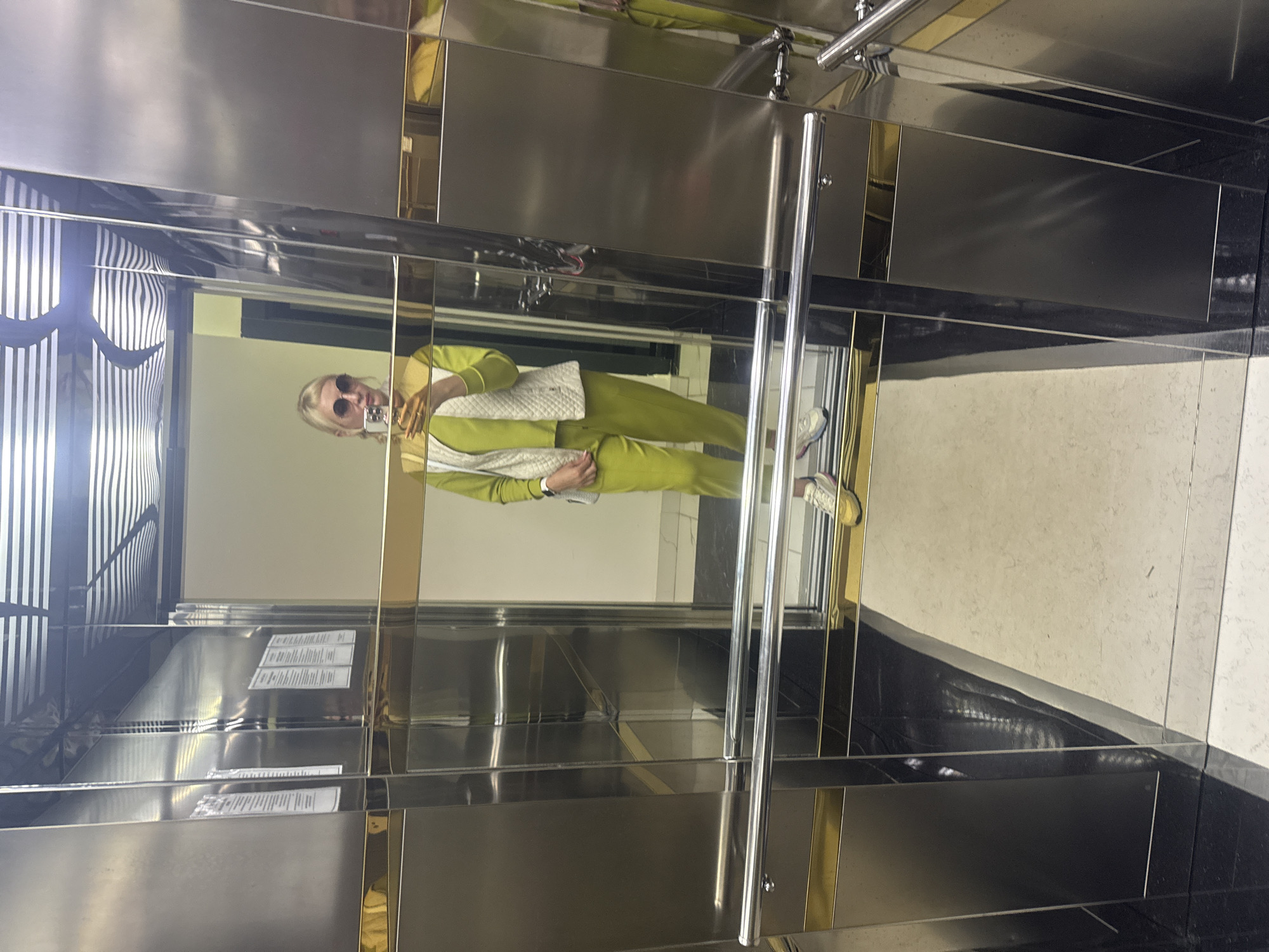 A person is visible in a mirror selfie taken inside a modern elevator. The individual has blonde hair and is wearing dark sunglasses. Their attire consists of a bright lime green tracksuit, featuring a long-sleeved top and matching pants, layered with a white quilted vest. White and pink sneakers are visible on their feet. The person is holding a phone in their right hand, positioned to capture the reflection.
The elevator's interior is characterized by highly reflective, possibly mirrored or polished chrome and gold-toned panels, creating a sleek and somewhat distorted environment due to multiple reflections. A silver handrail is visible on the right side of the frame. The elevator floor appears dark and reflective, possibly made of black marble or a similar polished material.
Reflections on the left side of the elevator show a patterned interior space with distinct vertical stripes. Several small, rectangular white papers with unreadable text, likely elevator notices or certifications, are reflected on the left panels. The overall lighting within the elevator appears bright. The image captures a casual moment, possibly during the day, with the person seemingly posing for the photograph.