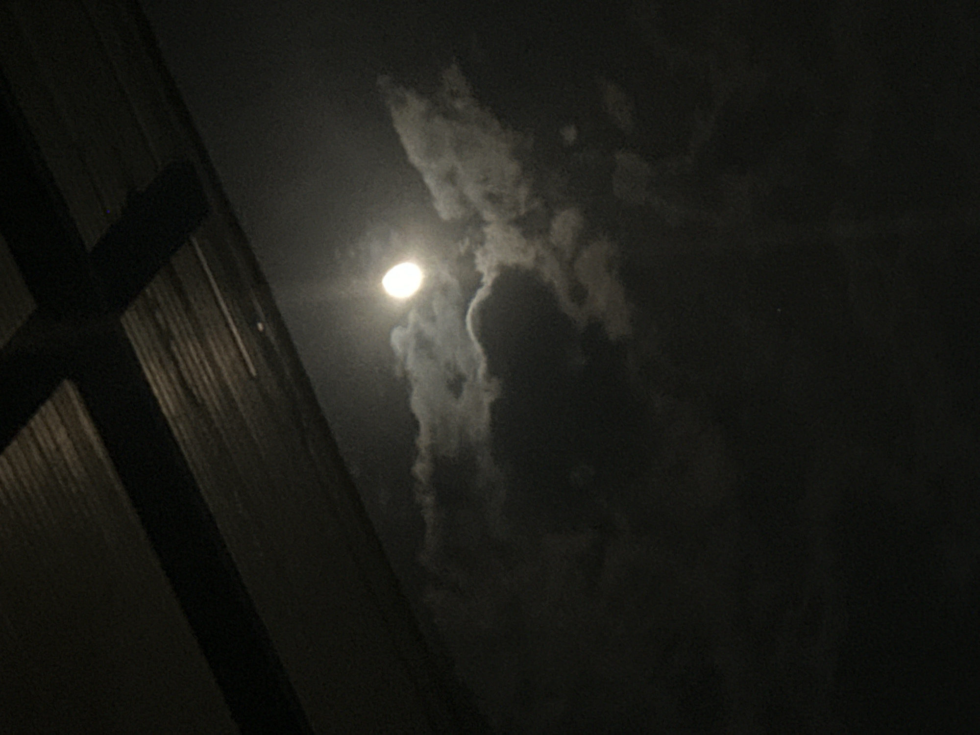 The image depicts a night scene with a bright moon partially obscured by clouds. To the left, there's a dark structure, likely a building, with vertical lines suggesting wood or metal siding. The sky is dark, with varying shades indicating cloud cover. The moon is a prominent, circular source of light, but its full shape is hidden behind a dense cloud formation. No people or specific objects are discernible beyond the building and clouds. The location is unknown.