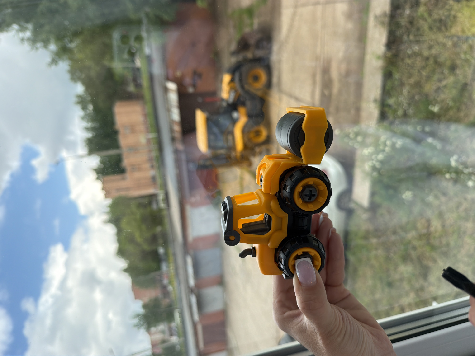 The image shows a person's hand holding a yellow and black toy construction vehicle, viewed through a window on a bright, clear day in Votkinsk, Russia. The hand, positioned on the right side of the frame, displays neat, light-colored nail polish. The toy, resembling a front-end loader or similar heavy machinery, is held upright.
Through the window, an outdoor scene unfolds. In the mid-ground, two real yellow construction vehicles are visible on an unpaved area, likely a construction site. One vehicle appears to be an excavator or large loader, while the other is partially obscured. Behind the construction area, lush green trees stand prominently, and in the distance, several brick buildings can be seen.
The window glass reflects a significant portion of a bright blue sky adorned with large, white, fluffy clouds, particularly across the upper half of the image. A faint, translucent reflection of what appears to be a smartphone camera module is also discernible in the upper left corner of the window. The scene depicts a moment of quiet observation or play, linking the toy in hand with the real-world activities outside. The time of day is clearly daytime, characterized by ample sunlight. No visible text is present in the image.