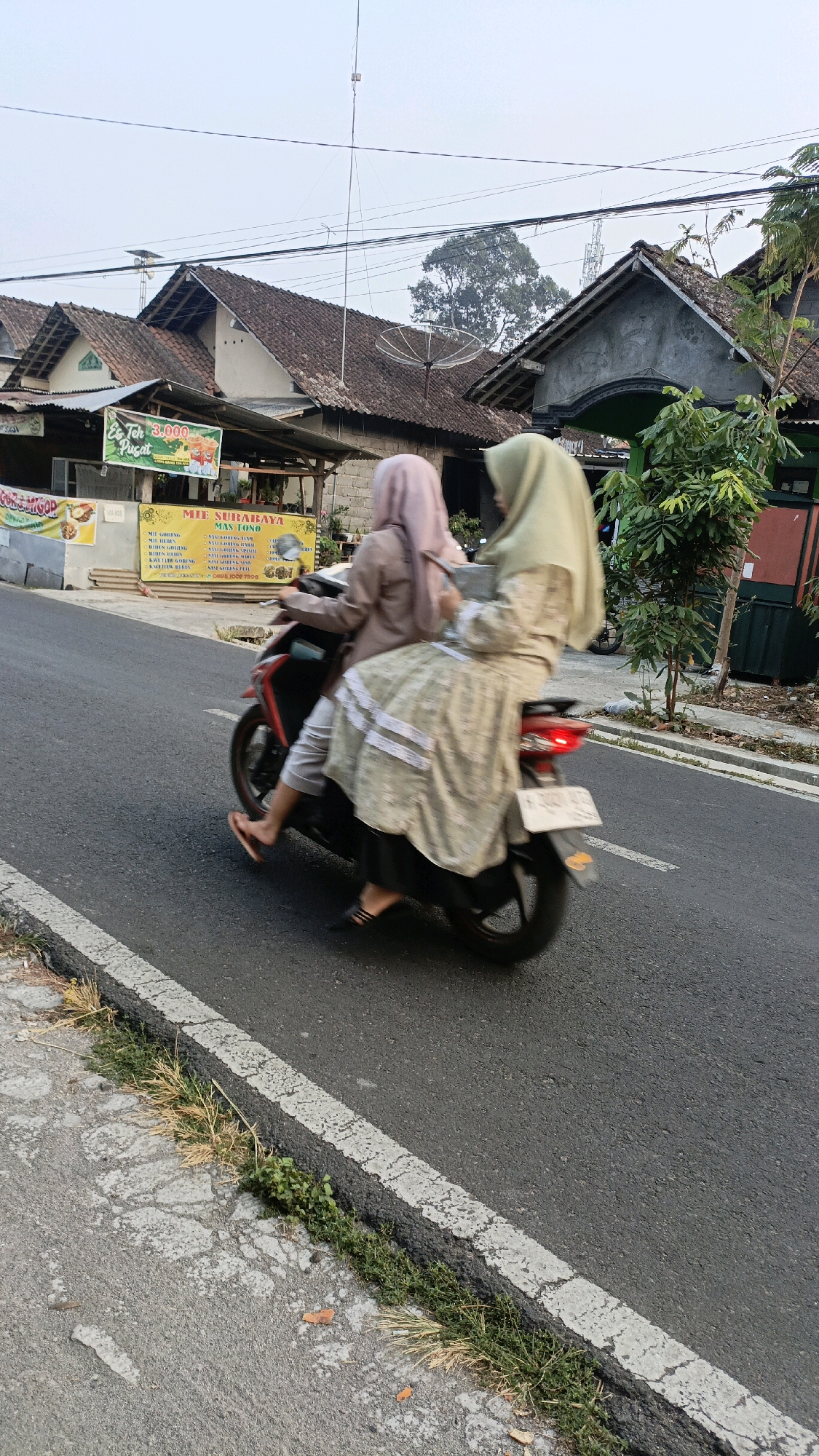 Two individuals, likely women wearing hijabs, are riding a red motorcycle on a paved road. The person in front is wearing a light brown jacket and white pants, while the person behind is wearing a light green hijab and a patterned dress. The motorcycle is moving from left to right, with motion blur indicating movement. The environment appears to be a street in Salatiga, Indonesia, with buildings and vegetation visible in the background. A sign in the background advertises "Mie Surabaya Mas Tono" and "Es Teh 3.000". The road has a white line marking the edge and is bordered by a sidewalk with grass and some debris. The sky is overcast.