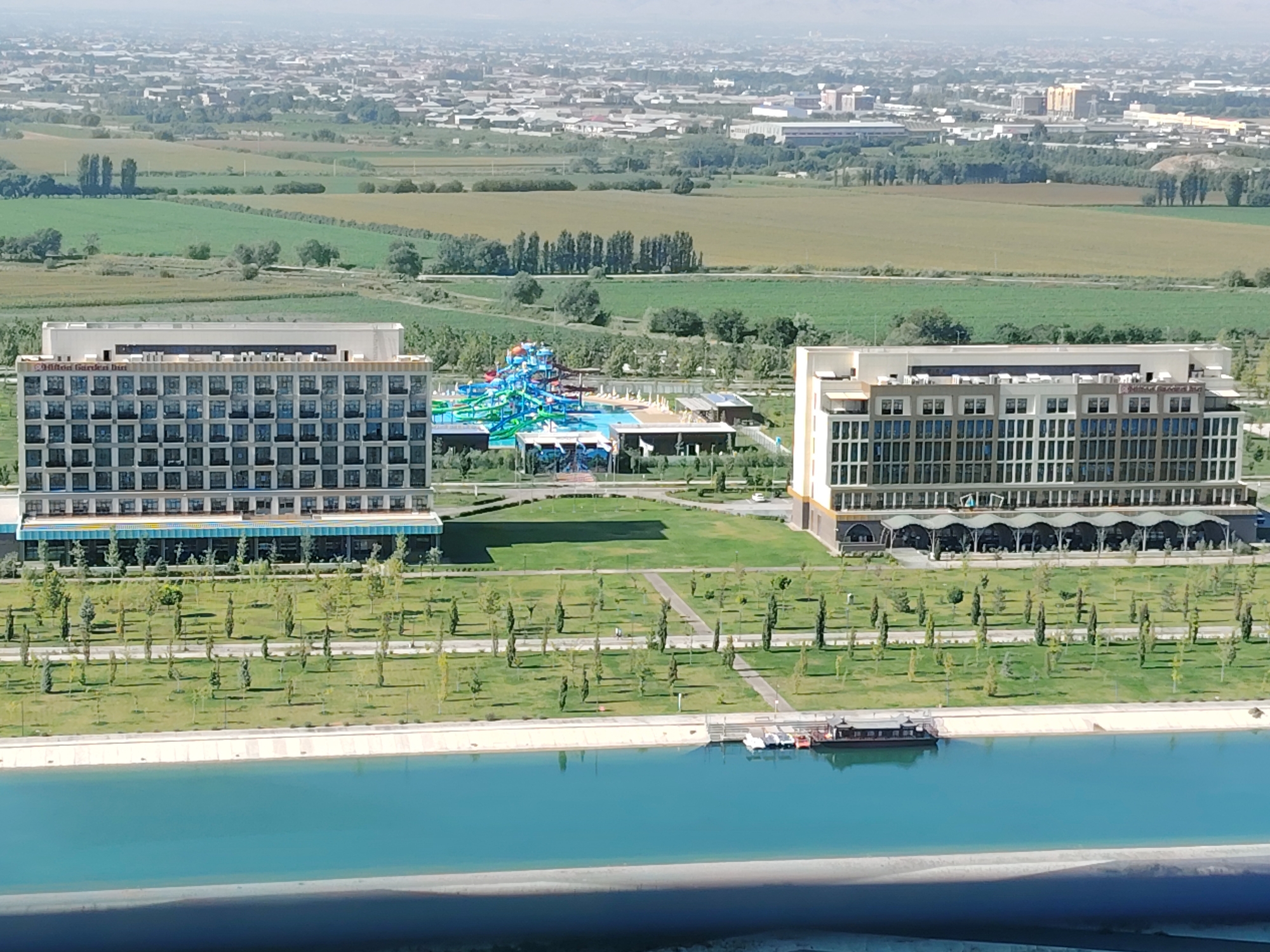 The image shows a panoramic view of a resort in Konigil, Uzbekistan, on a clear day. The foreground is dominated by a wide, bright turquoise canal. A dark brown, wooden boat with a covered top is docked on the right side of the canal, with several smaller white boats nearby.
Beyond the canal, a landscaped area with meticulously manicured green lawns and scattered trees leads up to two large, modern hotel buildings. The hotel on the left is a light beige structure with a grid of dark-framed windows. Above the windows, the text "Hilton Garden Inn" is visible. To the right of this hotel, a vibrant blue and green water park complex is situated, featuring water slides and pools.
The hotel on the right is a taller, beige building with a similar window pattern, but the windows are larger and more uniform. This building has a prominent arched canopy extending along its lower level, suggesting outdoor seating or a promenade. The background reveals a sprawling city with numerous buildings, extending towards a hazy horizon. Fields of varying shades of green and yellow are visible between the resort and the city, interspersed with rows of trees and some scattered smaller buildings. The overall impression is one of a modern resort complex set against a backdrop of urban development and agricultural land.
