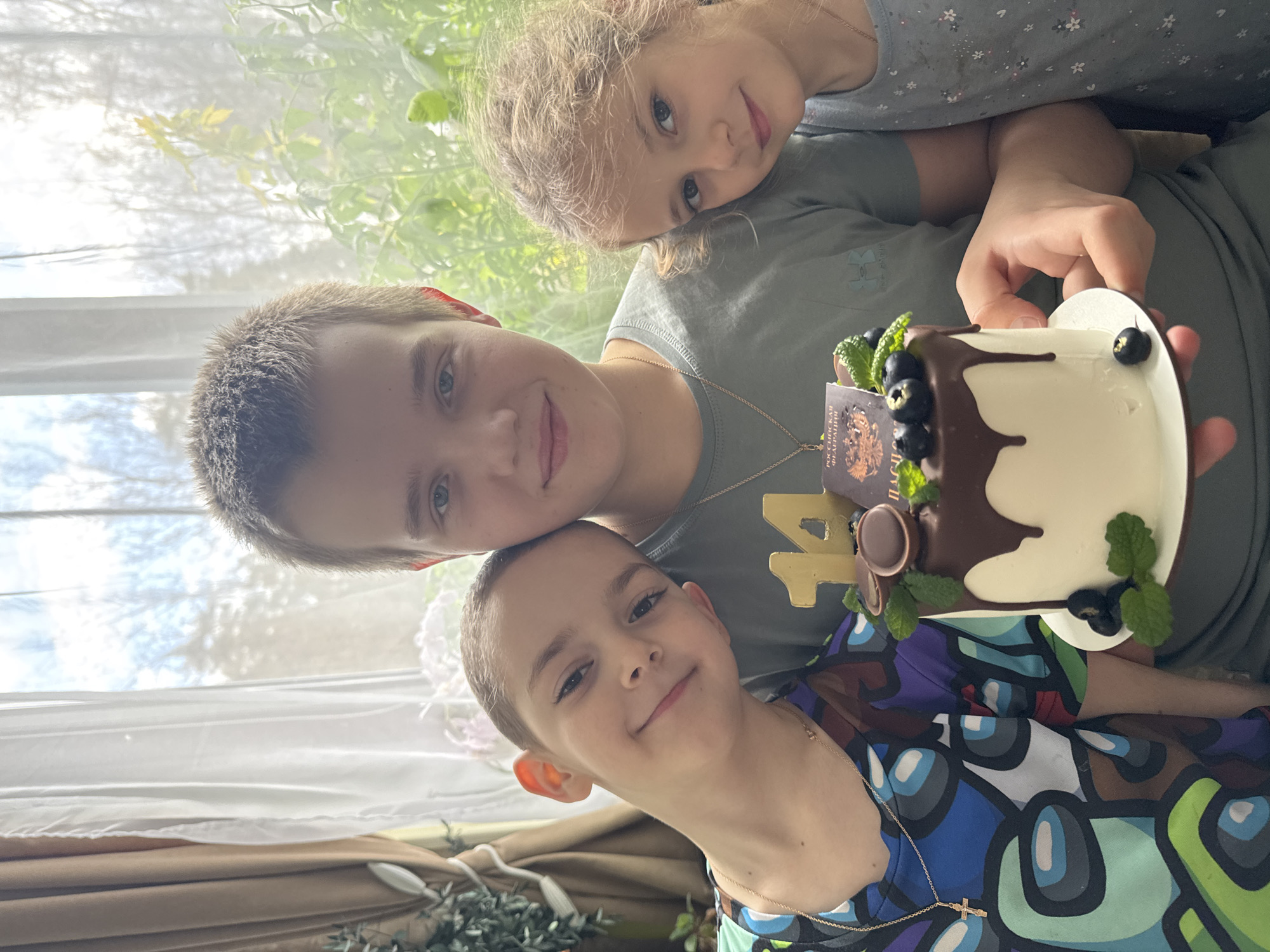 Three children are captured in a joyful, brightly lit indoor setting, likely celebrating a birthday or special occasion. The image is taken from a slightly upward angle, with natural light streaming in from a large window in the background, revealing blurry green foliage outside.
In the foreground, a young boy with short, light brown hair and blue eyes smiles warmly at the camera. He wears a colorful t-shirt adorned with cartoon-like characters, possibly from the game "Among Us," and a thin gold chain with a cross pendant. Slightly behind him and to his left, an older boy with short, light brown hair and blue eyes also smiles directly at the camera. He wears a dark grey or olive-green t-shirt with a gold chain. Resting his head gently on the older boy's shoulder is a young girl with blonde, curly hair and blue eyes, looking towards the camera with a sweet expression. She wears a lighter-colored top.
The older boy is holding a small, heart-shaped or possibly round white cake. The cake features chocolate ganache dripping down one side and is garnished with fresh blueberries and mint leaves. A golden number "14" is prominently displayed next to the cake, indicating the age being celebrated. A small, dark-colored rectangular decoration resembling a passport is also inserted into the cake, with the text "РОССИЙСКАЯ ФЕДЕРАЦИЯ" (Russian Federation) and a coat of arms clearly visible.
The overall mood is cheerful and celebratory, with all three children displaying happy expressions. The background shows white curtains and hints of indoor plants on the left, adding to the homey atmosphere. The bright lighting suggests it is daytime.