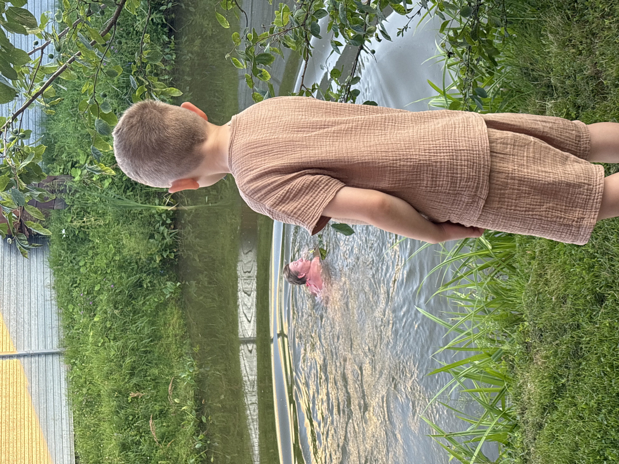 In a natural outdoor setting, two young children are partially submerged in a body of water. The scene appears to be during daylight hours, possibly late afternoon or early evening, given the soft lighting. One child, viewed from behind, stands near the edge of the water, wearing a textured, light brown garment. Their head is turned, looking towards the other child. This second child is further in the water, with only their head and shoulders visible above the surface. They are facing away from the camera, with their head tilted back, appearing to be enjoying the water.
The surrounding environment is lush and green, with abundant grass and foliage. Trees with leaves and small, green fruits are visible, their branches hanging over the water. The water itself is calm, with gentle ripples reflecting the surrounding light and greenery. The overall atmosphere suggests a relaxed, summery moment of outdoor play. There is no visible text in the image. The location context is identified as Дунилово, Russia.