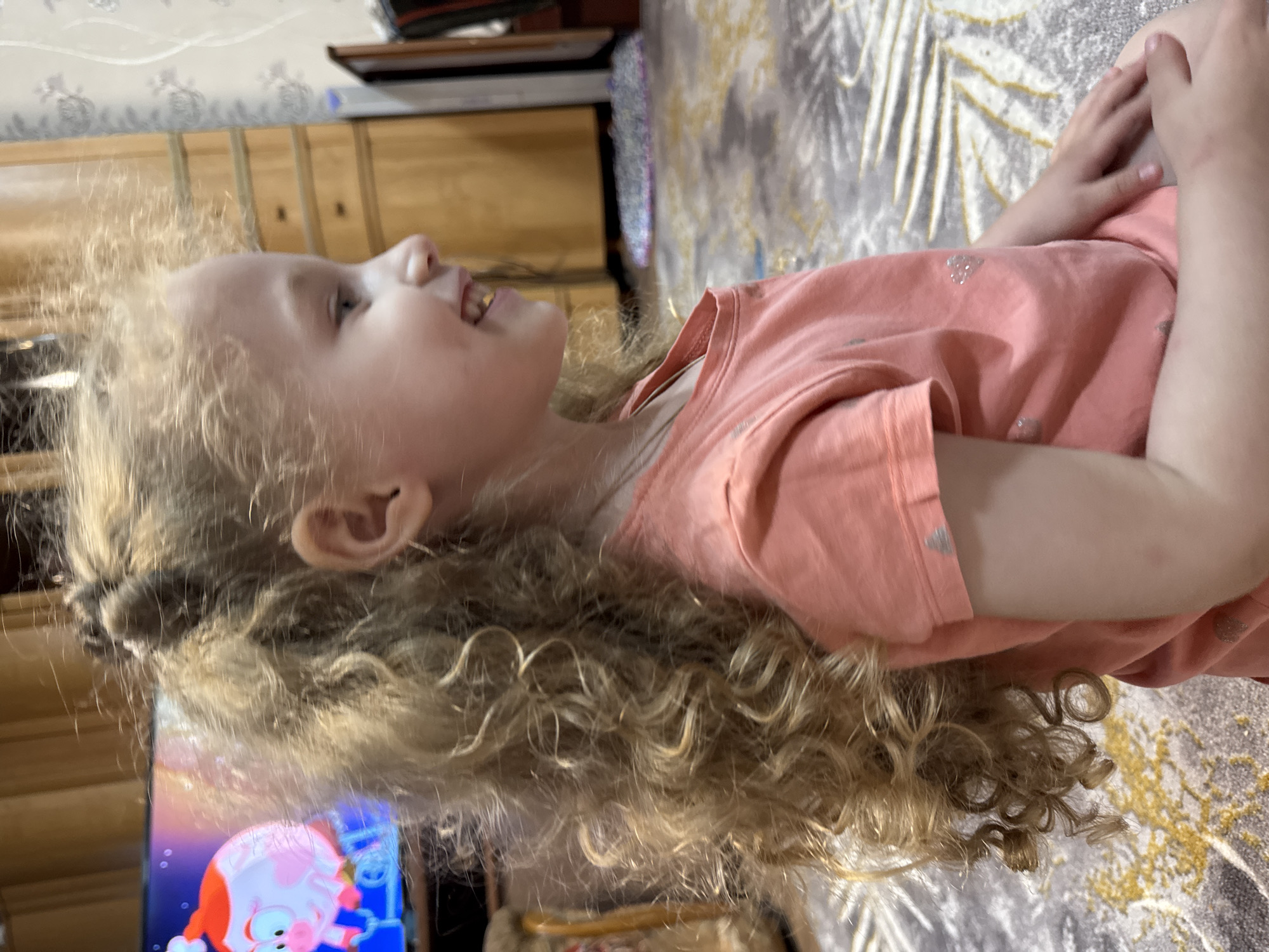 A young girl with curly blonde hair is lying down, facing to the left, wearing a pink shirt with silver patterns. The girl appears to be smiling. A television screen is visible in the bottom left, displaying cartoon imagery. A wooden dresser stands in the background, along with a gray and white patterned rug. The location is Nizhnyaya Gar', Russia.