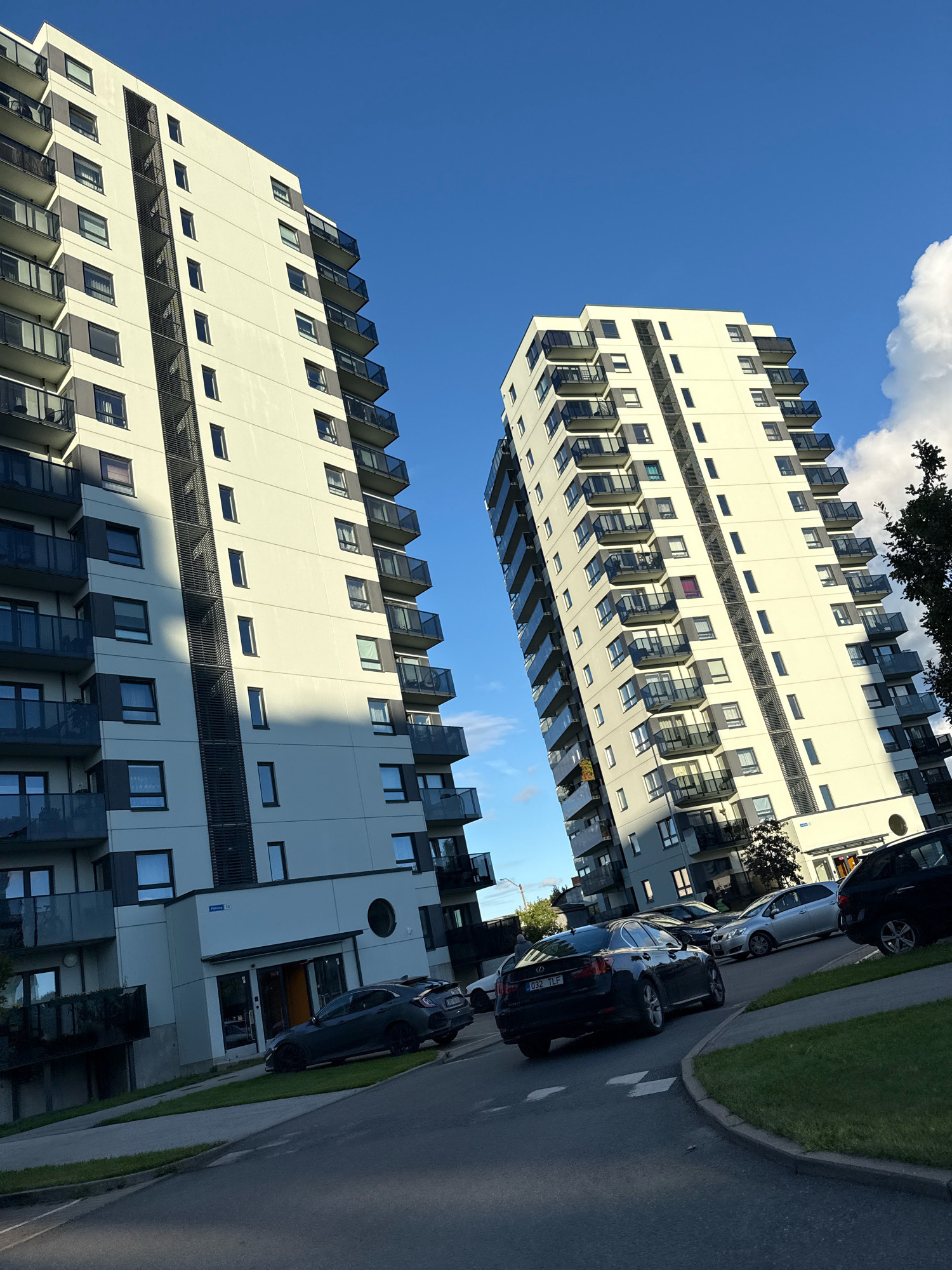 The image shows an outdoor view of two modern apartment buildings in Tallinn, Estonia, on a sunny day with a bright blue sky and scattered clouds. The buildings are tall, multi-story structures with light-colored facades, dark balconies with glass railings, and numerous windows. One building is positioned to the left of the frame, and the other is to the right, appearing slightly closer to the viewer and leaning. In front of the buildings, a street or driveway curves, and several cars are parked or driving. A dark grey sedan is parked on the right side of the driveway, facing away from the camera. To its left, a silver hatchback is visible. Further in the background, another dark car is parked. The ground in front of the buildings is a mix of paved surfaces and green grass. A sign on the building on the right reads "Fabriku 12". The overall scene depicts a residential area in an urban setting.
