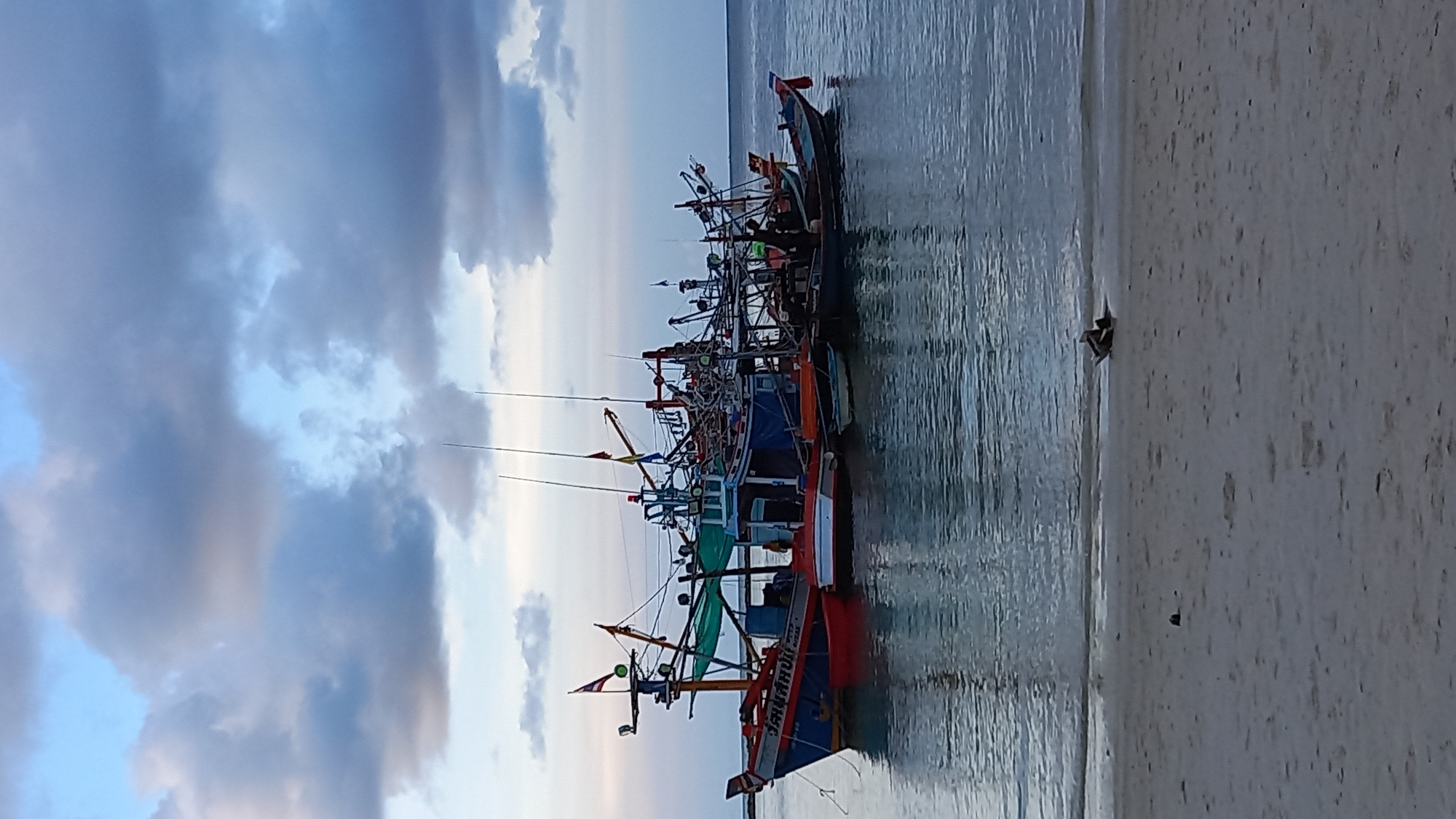 The image shows a group of fishing boats moored in calm water near a sandy beach. The boats are of various sizes and colors, with some displaying flags and antennae. The sky is partly cloudy with a mix of blue and gray tones. In the foreground, the sandy beach has some debris, and a person appears to be lying down or resting on a small raft or float near the water's edge. The scene appears to be set in a coastal area, likely in ตำบลเกาะพะงัน, Thailand, given the presence of numerous fishing vessels.