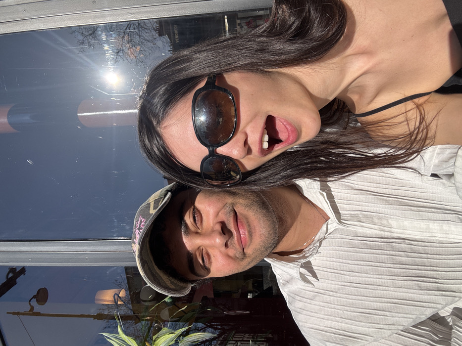 The image depicts two individuals, a male and a female, positioned closely together in a bright outdoor setting. The male, on the left, wears a camouflage-patterned baseball cap and a white, vertically pleated button-down shirt, looking directly at the viewer with a slight smile. The female, on the right, wears large, dark-framed sunglasses and a black spaghetti-strap top, with her mouth wide open in an expression of surprise or excitement.
Behind them, a large glass window reflects elements of the exterior environment. Visible in these reflections are a bright sun, bare tree branches, a distinctive black street lamp with a conical shade, and portions of a light-colored building. The French word "VENDREDI" (Friday) is clearly reflected in white text on a dark background on the right side of the glass. This textual detail, combined with the typical Parisian street architecture and lamp design visible in the reflection, places the scene in Paris, France. The lighting suggests a sunny day.