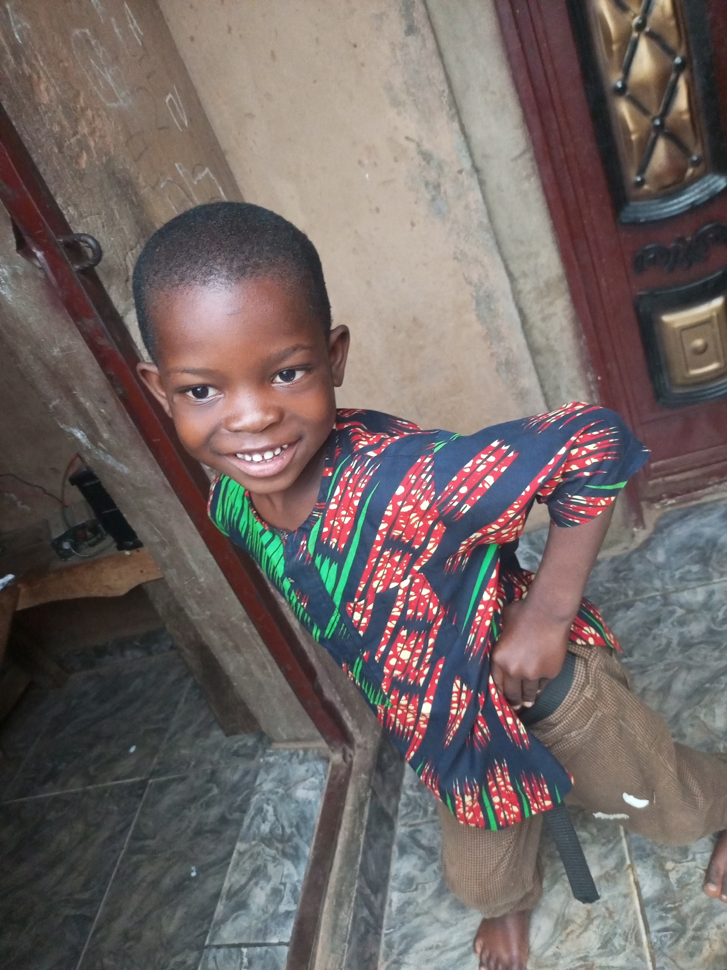 A high-angle full shot captures a young Black boy standing in what appears to be an entryway with a tiled floor and a wooden doorframe. The boy, with short black hair and dark eyes, faces the camera with a wide, toothy grin. He is wearing a patterned shirt with red, yellow, and green accents on a black background, and brown checkered pants with a black belt. His left arm is bent, and his hand is near his waist. To his left, a dark metal railing is visible, and further back, a piece of electronic equipment is mounted on a wooden surface. The wall behind him is textured and light-colored, with some faint markings. To his right, a dark red wooden door with decorative gold accents is partially visible. The floor is covered in gray and black marbled tiles.