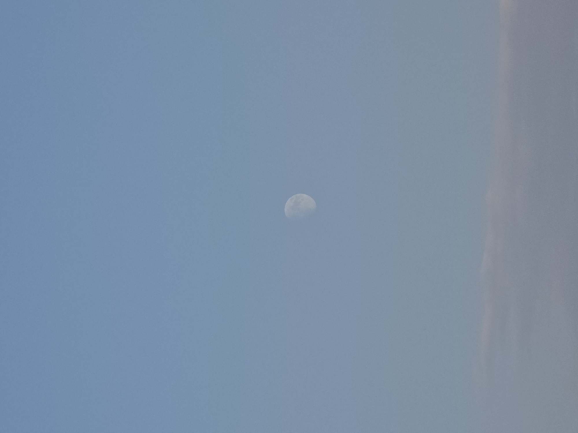 The image captures a clear blue sky with a visible crescent moon positioned slightly off-center to the left. Faint clouds are present at the bottom of the frame, appearing as wispy horizontal bands. The scene depicts a daytime sky, likely in Obrochishte, Bulgaria, characterized by its expansive and serene atmosphere. There are no people or specific objects depicted beyond the celestial bodies.