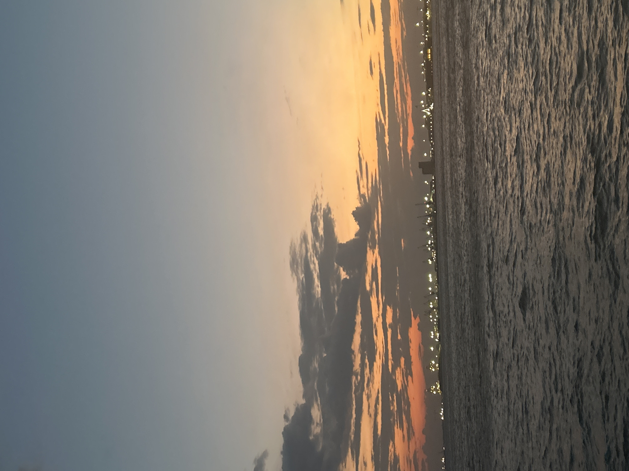 The image captures a twilight scene over the sea, with the city skyline of Thessaloniki visible in the distance. The sky transitions from a pale blue at the top to a vibrant orange and red near the horizon, where scattered clouds add texture to the sunset. The sea in the foreground is a deep, choppy gray, reflecting the dimming light. The distant city skyline is illuminated by numerous lights, suggesting a port or industrial area, with several cranes and structures identifiable. There are no people or animals visible in the frame. The overall impression is one of a serene, albeit industrial, coastal landscape at dusk.