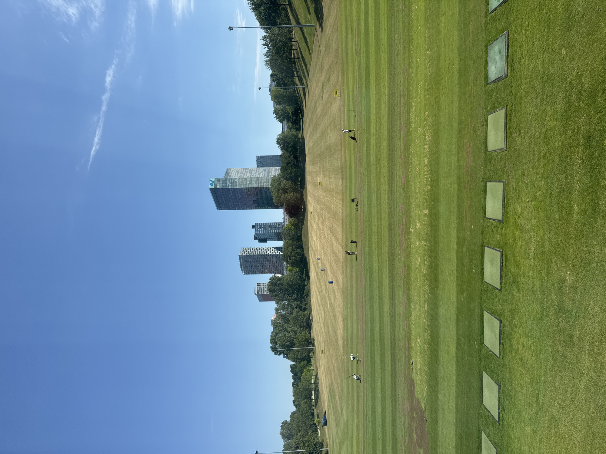 This image captures a sunny day at the Vienna Woods Golf Course, situated in Vienna, Austria. The foreground features a series of square green mats, indicative of a driving range, set against a backdrop of neatly mowed green grass. Beyond the mats, the expansive golf course unfolds with undulating terrain, dotted with several people practicing their golf swings. Yellow markers with numbers like "50" are visible on the course, denoting distances. Further in the distance, a line of trees separates the golf course from a cluster of modern high-rise buildings, including a prominent one with a blue logo. The sky is a clear, vibrant blue with a few wispy clouds. The overall scene conveys a sense of leisure and outdoor activity within an urban environment.