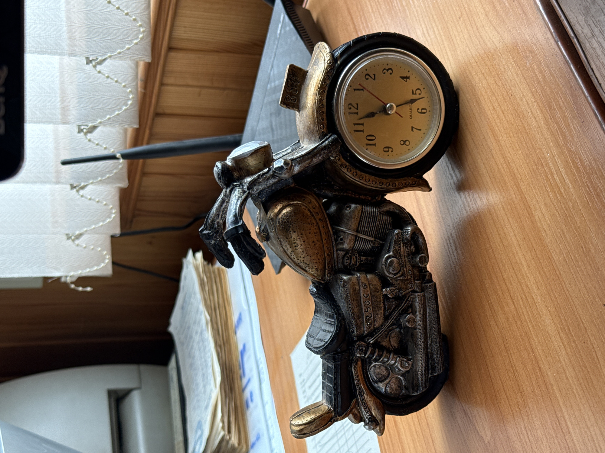 A close-up, eye-level shot shows a detailed, decorative clock in the shape of a motorcycle. The motorcycle is made of metal with a bronze-like finish and black accents. The clock face is integrated into the front wheel, featuring a yellow dial with black numerals and hands. The motorcycle is positioned on a light brown wooden desk. In the background, a stack of papers is visible on the left, and a light-colored beaded curtain hangs in front of a wooden wall and window on the right. A black antenna is also visible behind the motorcycle.