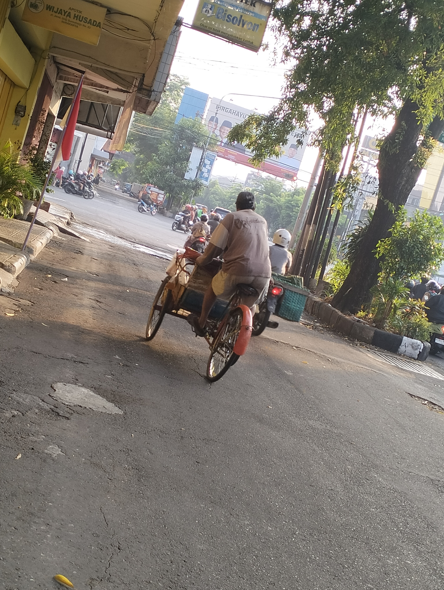 The image captures a street scene in Surakarta, Indonesia, during daylight. In the foreground, a person is riding a bicycle with a sidecar, commonly known as a becak. The rider is wearing a light-colored shirt and shorts. Behind the rider, the sidecar is visible, appearing to be made of woven material and carrying some items. The becak is moving on an asphalt road that shows signs of wear and tear, with visible cracks and patches.
In the background, other vehicles, including motorcycles, are also present on the road, suggesting moderate traffic. The roadside features some vegetation, including trees and potted plants. Buildings line the street, with one prominently displaying signage that reads "APOTIK WIJAYA HUSADA" and "Jl. Jend. Urip Sumoharjo No. 63 SURAKARTA". Further down the street, there are more buildings and advertisements. The overall atmosphere suggests a typical urban environment with everyday activities taking place. The sunlight indicates it is likely morning or late afternoon, casting shadows on the road.