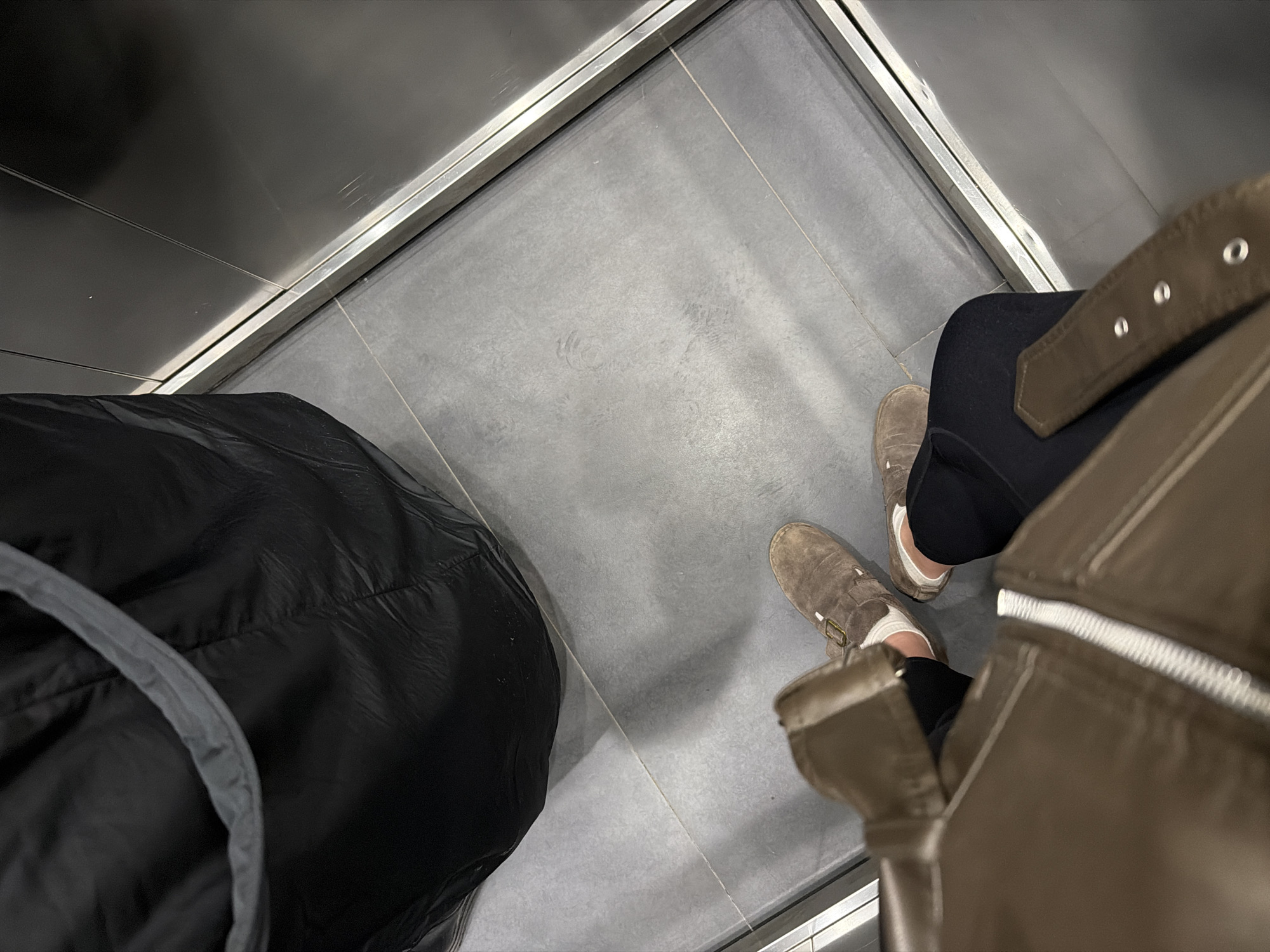 The image depicts an overhead perspective inside what appears to be an elevator. The elevator's walls are a reflective grey material, and the floor consists of large grey tiles. The view captures the lower portion of a person wearing dark pants, brown suede shoes with white socks, and a brown jacket. Beside the person is a large black bag with a grey strap. The scene is contained within the small square space of the elevator. It is impossible to determine from the image if the location is in Moscow, Russia.