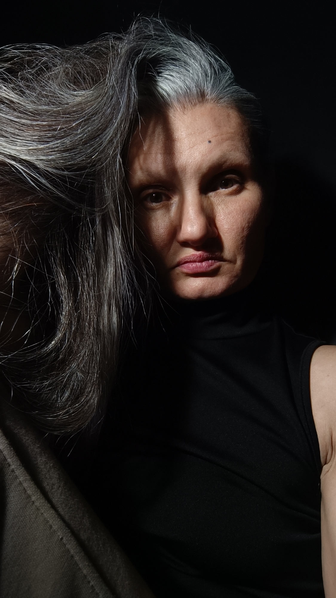 A medium close-up shot depicts a mature woman with gray hair, one side of which obscures part of her face. The woman is wearing a black sleeveless top. The lighting is dramatic, casting deep shadows across her face. The background is dark and indistinguishable. There is no evidence to indicate that the location is Lermontov, Russia.
