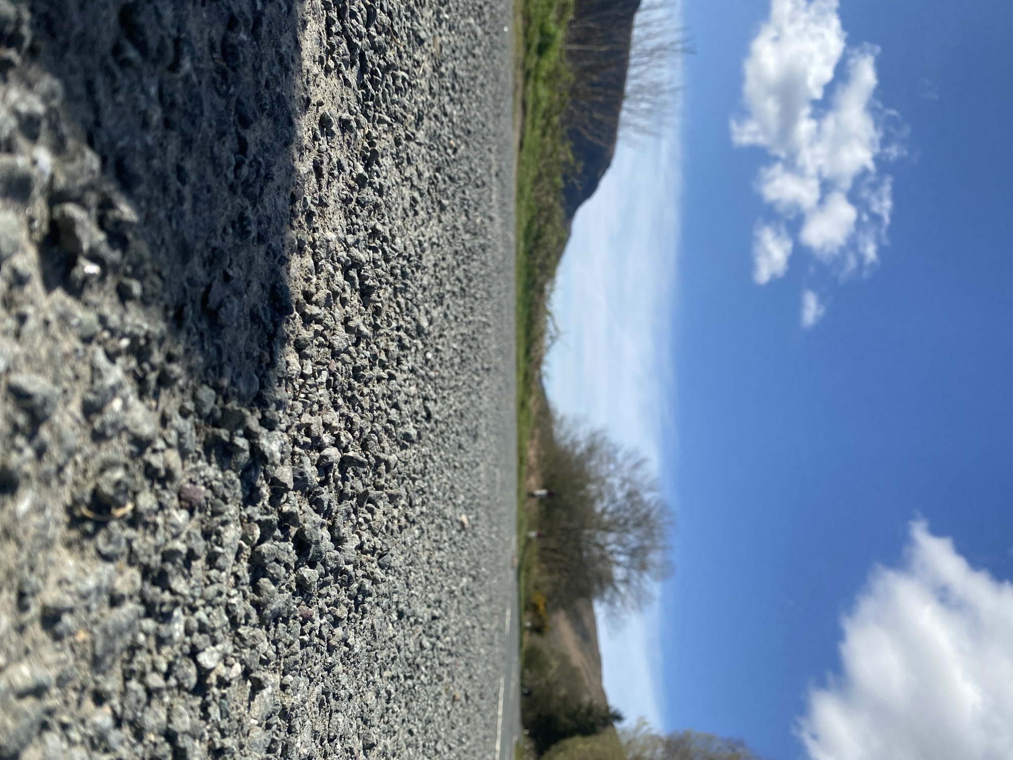 The image depicts a low-angle, perspective view of an asphalt road or pavement surface, textured with small aggregate stones, in Llangynog, United Kingdom. The road occupies the left half of the frame. The right half shows a landscape comprising grassy areas, shrubs, trees, and gently sloping hills. A blue sky with scattered white cumulus clouds fills the upper portion of the scene.