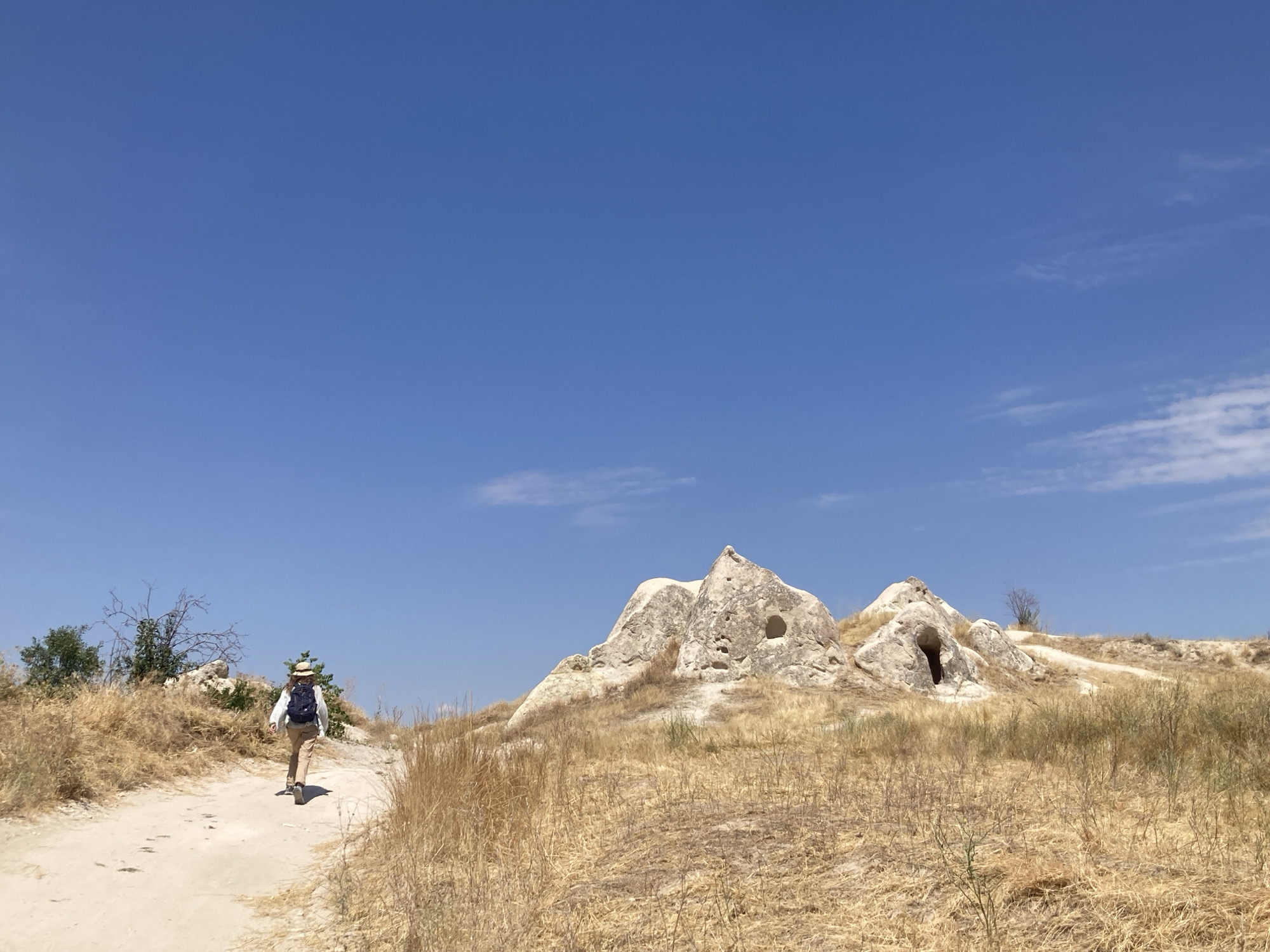 A person wearing a hat and backpack walks along a dirt path in a dry, grassy landscape. The path curves to the left, leading away from the viewer. To the right of the path, there are unusual rock formations with caves or openings carved into them. The sky is a clear, bright blue with a few wispy clouds. The scene appears to be set in Göreme, Turkey, a region known for its unique geological formations and ancient cave dwellings.