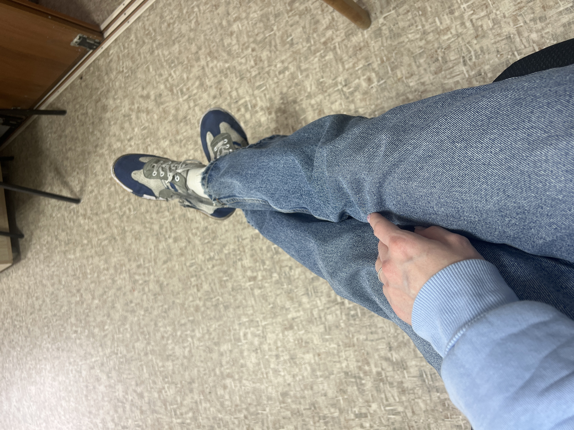 A view from above shows a person sitting in a chair, wearing blue jeans, a light blue sweater, and blue and gray sneakers. The person's right hand is resting on their right leg. The floor is a light gray and beige patterned linoleum. The location cannot be determined to be Chaikovsky, Russia.