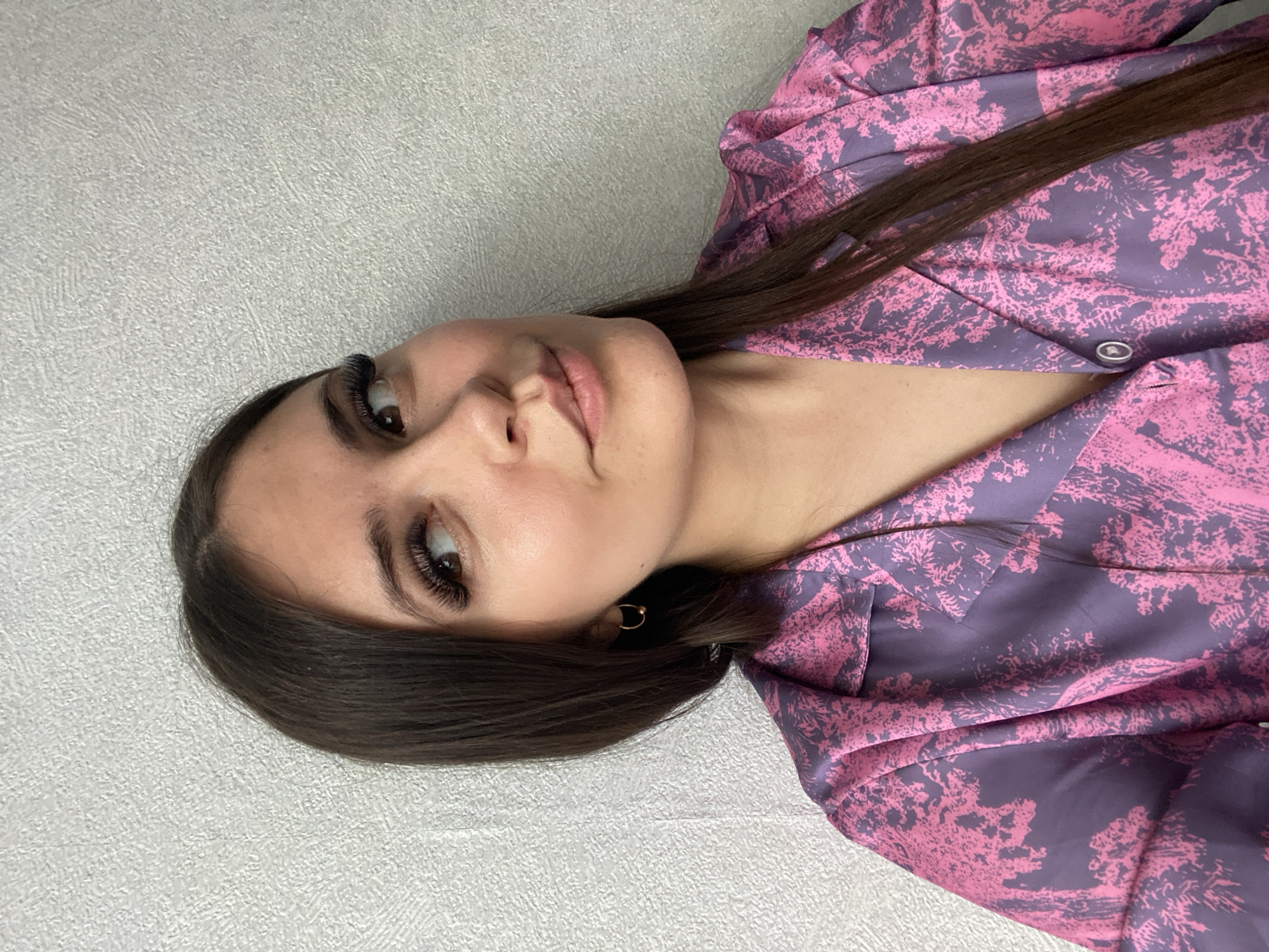 A young woman with long, dark brown hair is pictured indoors, against a light grey wall. She is wearing a patterned blouse with pink and purple hues. The photo focuses on her face, featuring prominent eyelashes and light makeup. She is wearing a small, gold earring in her left ear. There is no readily available information within the image to confirm the location in Chaikovsky, Russia.
