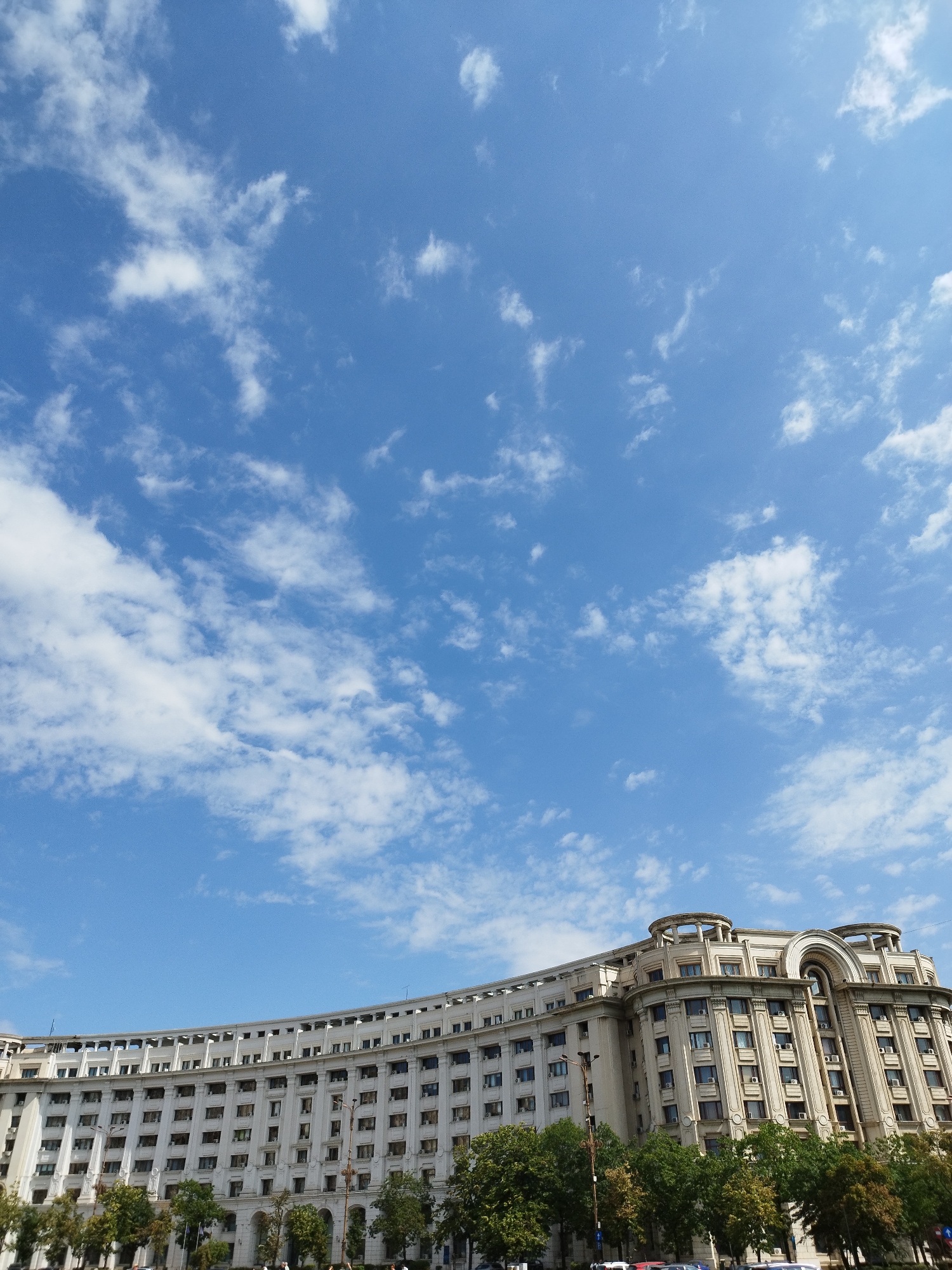 The image shows the Palace of the Parliament, also known as the People's House, in Bucharest, Romania. It is the second-largest administrative building in the world and the heaviest building in the world. The building is a monumental structure of socialist realism architecture, characterized by its imposing size, symmetrical design, and extensive use of marble and gold. The image captures a low-angle view of the building under a bright blue sky with scattered white clouds. Trees line the foreground, and a few streetlights are visible. The architecture is ornate, with numerous windows, columns, and decorative elements. The scale of the building is emphasized by the vast expanse of the sky above.