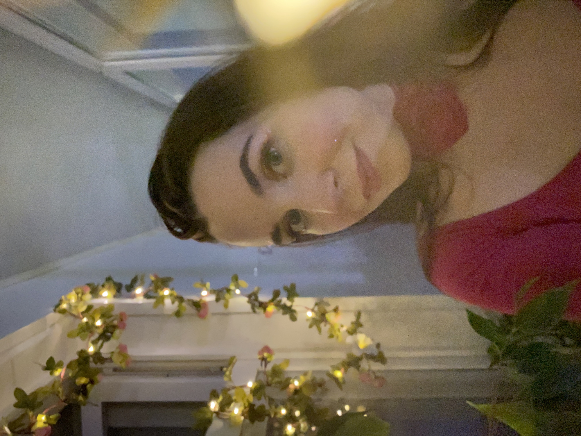 The image displays a close-up, indoor portrait of a female. The specific location in the city of Chaikovsky, Russia, cannot be determined from the visual evidence within the image.
The subject is a woman with long, dark brown hair and light-colored eyes, looking directly at the camera with a subtle smile. She has a fair complexion and appears to be wearing makeup, including eyeliner. A small, shiny, metallic-appearing detail is affixed to her lower lip, slightly to the left. Her attire consists of a vibrant pink or fuchsia-colored top with a v-neckline. Around her neck, a red fabric accessory, possibly a bow or flower, is visible, positioned slightly to the right of her central axis.
To the left of the woman, a decorative garland is strung vertically, featuring small, warm white LED lights intertwined with artificial green leaves and pale pink flowers. This garland appears to be attached to a window frame, with a dark area, likely a window pane, visible beyond it. In the lower-left foreground, green foliage from an indoor plant is partially visible. The upper portion of the background shows a plain, light-colored wall or ceiling. A strong, warm light source, originating from the right side and outside the frame, produces a prominent lens flare and bokeh effect, partially obscuring the right side of the woman's head and the background. The lighting conditions suggest an evening or night setting.
