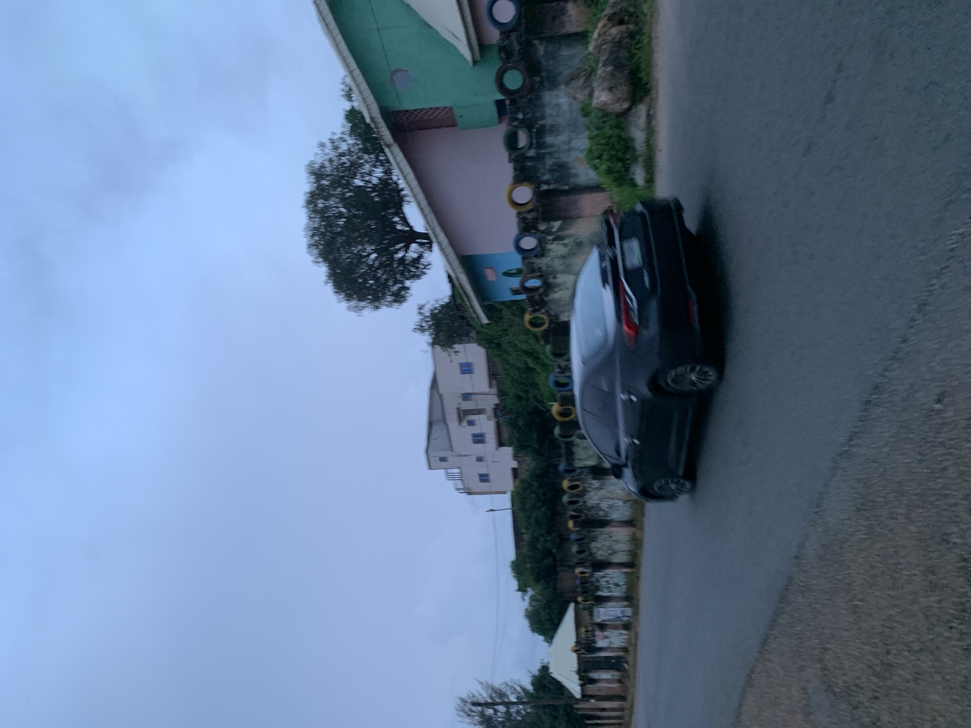 A dark-colored sedan drives along a paved road in Jos, Nigeria. The car is angled away from the viewer, with its rear left side most prominent. The road surface is a dark asphalt, with a lighter colored gravel shoulder on the left.
Behind the road, a low, weathered concrete wall runs horizontally, decorated with a series of colorful, repurposed tire sections. Beyond the wall, the terrain slopes upwards, covered with lush green vegetation and scattered rocks.
Several buildings are visible on the elevated ground. To the left, a multi-story building with a light-colored facade and blue windows stands against a pale, overcast sky. Further to the right, a row of smaller, brightly colored structures is visible, including a pink building and a vibrant green building, each with white trim and visible windows. A large, leafy tree with a dark silhouette is prominent between these buildings.
The overall lighting suggests an overcast day, possibly early morning or late afternoon, with diffuse light and muted colors. The sky is a pale blue, with some wispy cloud formations. The atmosphere is calm and still.