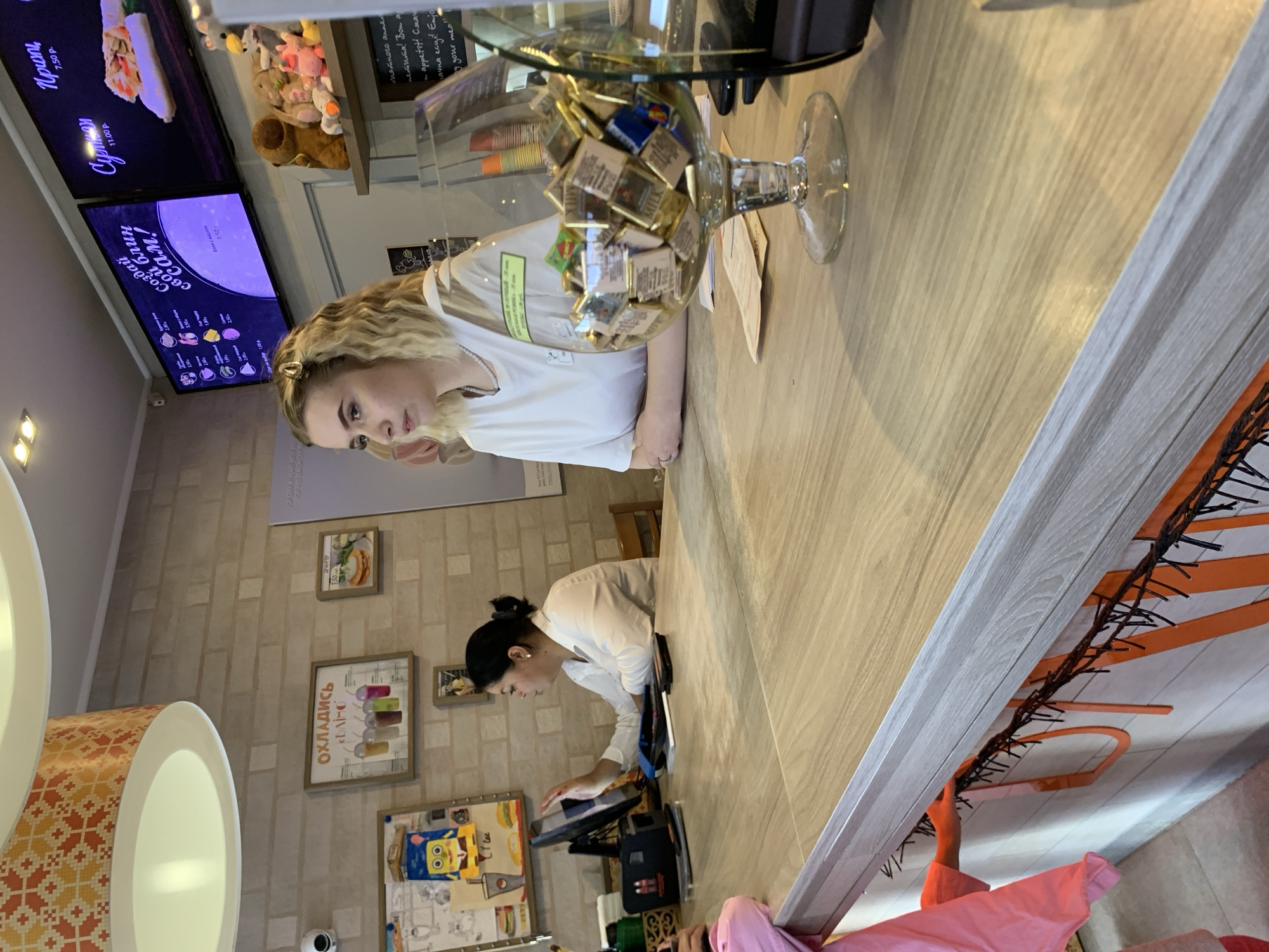 A young woman with blonde curly hair, wearing a white t-shirt and pearl necklace, stands behind a wooden counter in what appears to be a cafe or fast-food establishment. She is looking to her left. To her left, another woman with dark hair pulled back, wearing a white long-sleeved shirt, is operating a touchscreen device. In the foreground, a large wine glass filled with small, individually wrapped chocolates sits on the counter. Behind the counter, shelves display stuffed animals and framed pictures with food imagery. Digital menu boards are visible on the wall behind the counter. The environment is brightly lit, with decorative lighting fixtures visible in the upper portion of the image. Orange string lights are draped along the front of the counter. The establishments' location in Sluck, Belarus, is not discernible from the visual information provided in the image.