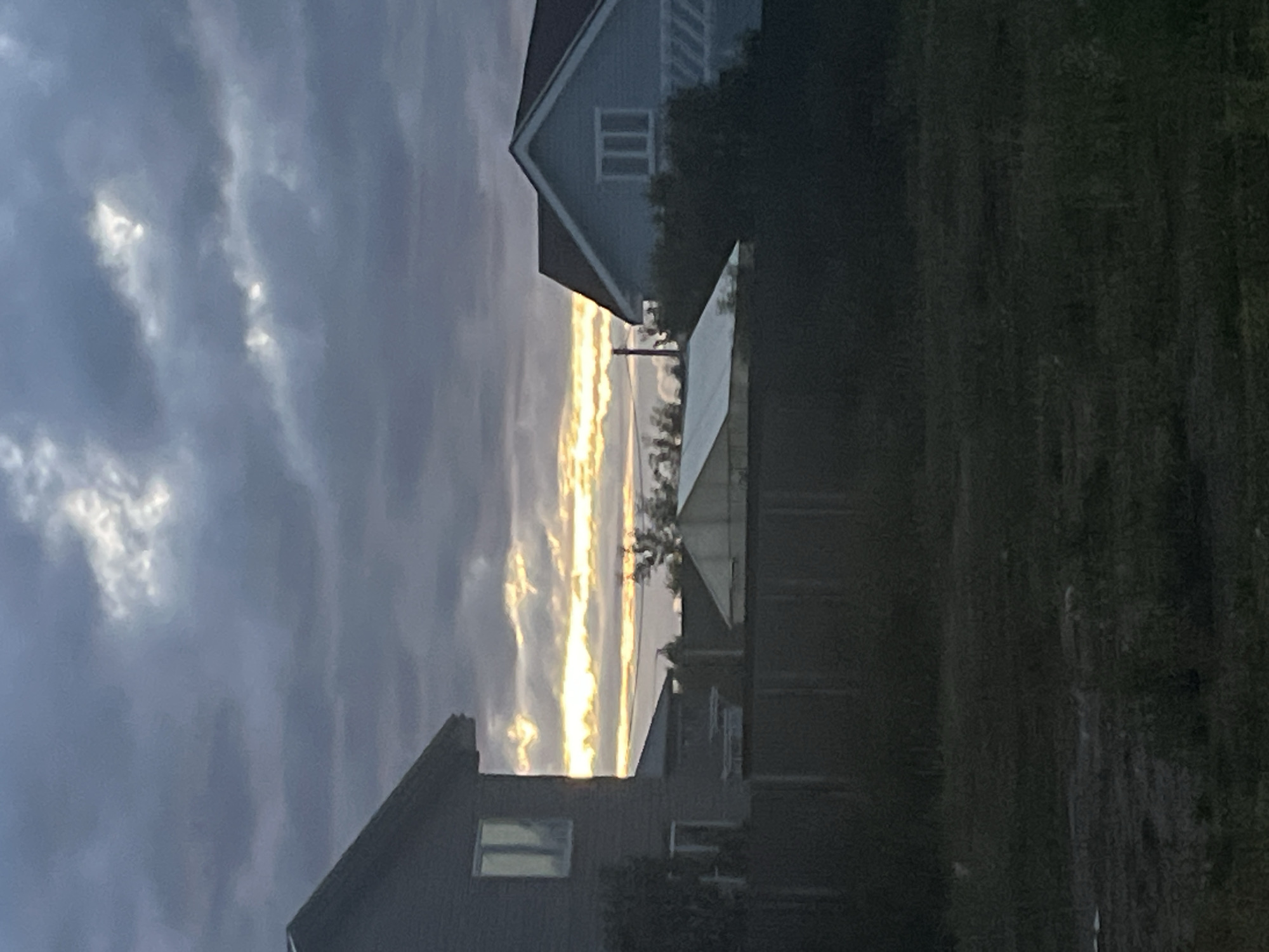 The image depicts a rural landscape at dusk, possibly in Vladimirovka, Russia, with a focus on residential structures and the sky. Two houses are visible: a darker, two-story house on the left with a prominent gabled roof and a window reflecting the sky, and a lighter blue, single-story house on the right with a red roof and a multi-paned window. Between the houses, a greenhouse-like structure with a white, sloped roof is situated. A wooden fence runs horizontally across the mid-ground, separating the properties from a dark, grassy foreground. The sky is dominated by dramatic, layered clouds, with a striking band of yellow and orange light breaking through, suggesting a sunset or sunrise. The overall atmosphere is subdued, with muted colors and a sense of approaching night. No people or animals are present in the scene.