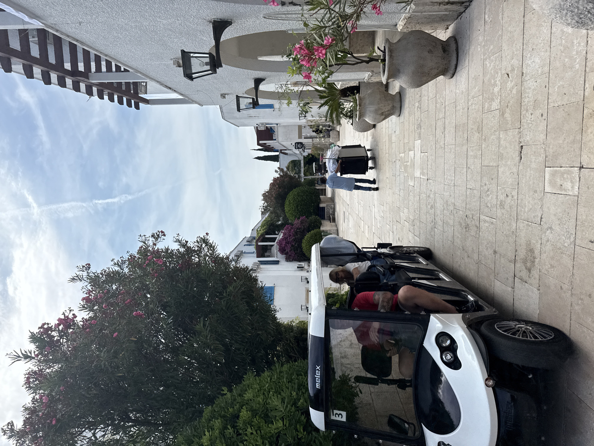 An exterior, eye-level, wide shot shows a white and black Melex golf cart with a "3" on its side, parked on a stone-paved pathway. A man in a red shirt is driving the cart, and a woman wearing glasses and a white top is seated in the back. The cart is positioned on the left side of the frame. To the right of the pathway, a white building with arched openings and lanterns is visible. Several large, gray planters with pink flowering plants are placed along the building's facade. In the background, two individuals are pushing a trolley laden with white linens. The scene is set in a narrow street lined with trees and buildings with a Mediterranean architectural style, characteristic of Budva, Montenegro. The sky is partly cloudy with a light trail of a plane.