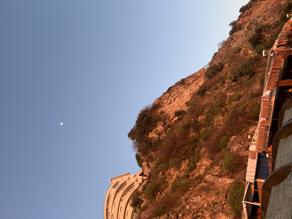 The image depicts a coastal scene in Rafailovići, Montenegro, under golden hour lighting. The upper two-thirds of the frame are dominated by a clear, light blue sky with a distinct crescent moon positioned in the upper left quadrant.
The lower two-thirds feature a steep, rocky cliff face, illuminated with warm orange and red tones from the sun. The cliff exhibits visible stratification and is sparsely covered with green and brown vegetation, including shrubs and small trees.
Perched atop the cliff on the left side is a multi-story building, likely a hotel or residential complex, characterized by light-colored facades and curved balconies. Further right on the cliff face, a set of white or light-colored stairs ascends vertically.
At the base of the cliff, in the foreground and lower mid-ground, a series of low-rise structures with white awnings and thatched roofs are visible. These structures appear to be beachside establishments, possibly restaurants or bars, with some featuring blue panels. In the immediate foreground on the left, portions of large, rounded, wooden or thatched beach umbrellas are visible. No individuals are present in the frame.