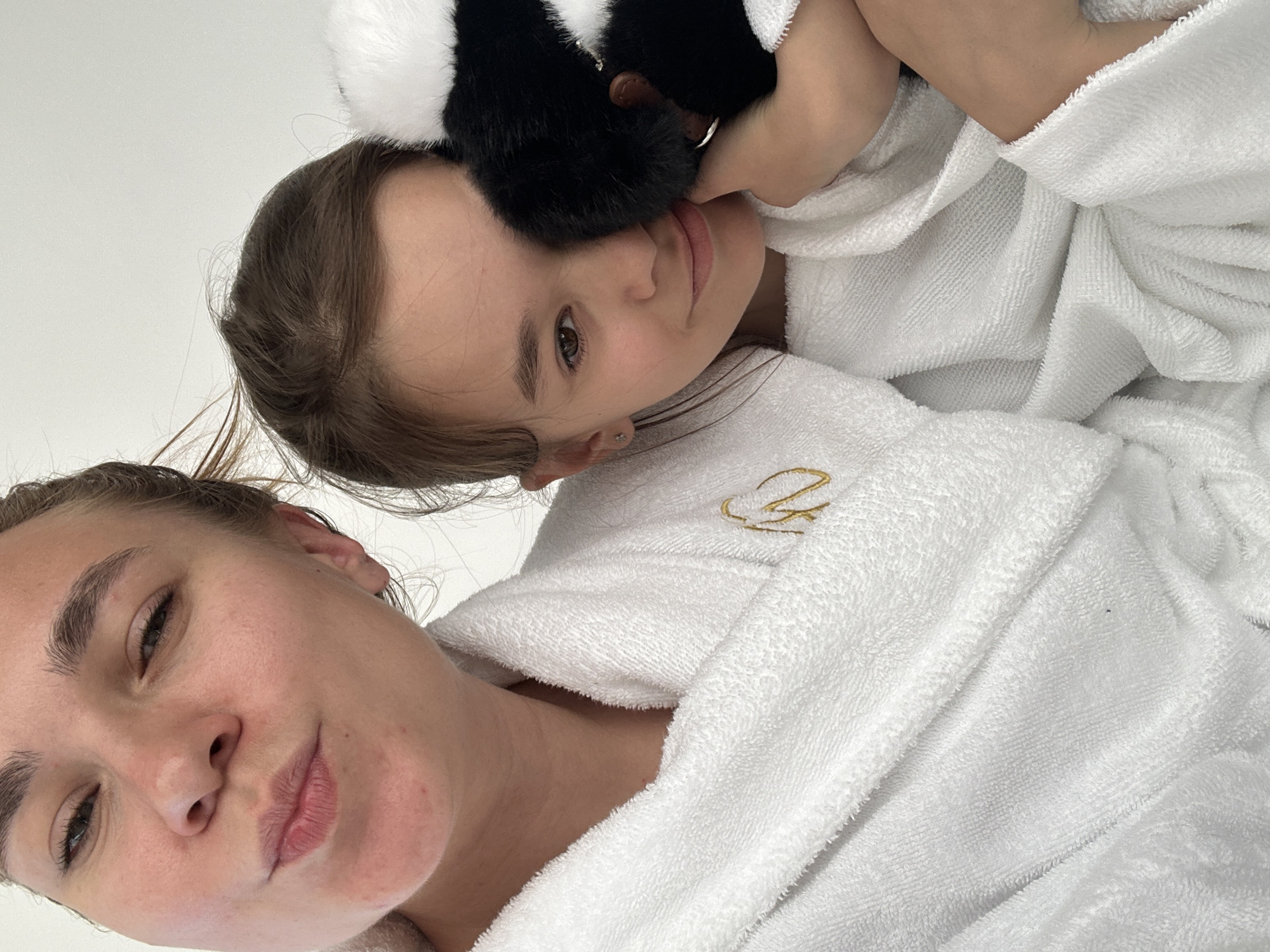 Two individuals, likely an adult and a child, are pictured in a close-up shot, both wearing white terry cloth robes. The adult, on the left, is looking towards the camera with their lips pursed. The child, on the right, is looking slightly away from the camera and holding a black and white plush toy. The robes appear to be soft and absorbent, suitable for spa or bath use. The background is a plain white wall, suggesting an indoor setting. The image quality is high, with clear focus and good lighting. The scene does not provide any specific clues about the location in Terskol, Russia.