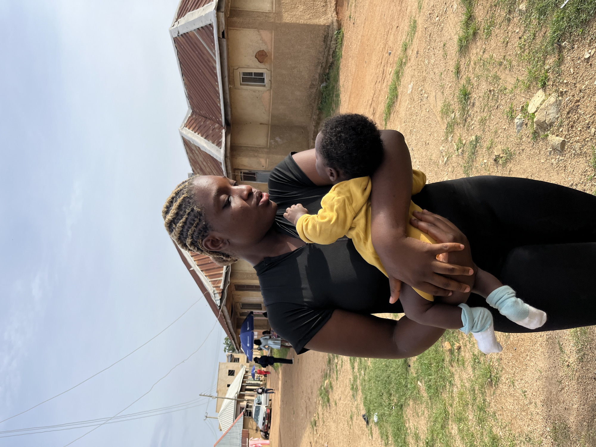 A young woman, likely of Nigerian descent given the location context of Rafin Par, Nigeria, holds a baby wrapped in a yellow garment. The woman has her hair in braided cornrows and is wearing a black top. The baby is asleep and is wearing light blue and white socks. They are outdoors on a dirt path with sparse green grass. In the background, there are buildings with corrugated metal roofs and some people walking around. The sky is overcast. The scene appears to be during daylight hours. The overall atmosphere seems peaceful, with the woman cradling the baby affectionately. There is no visible text in the image.