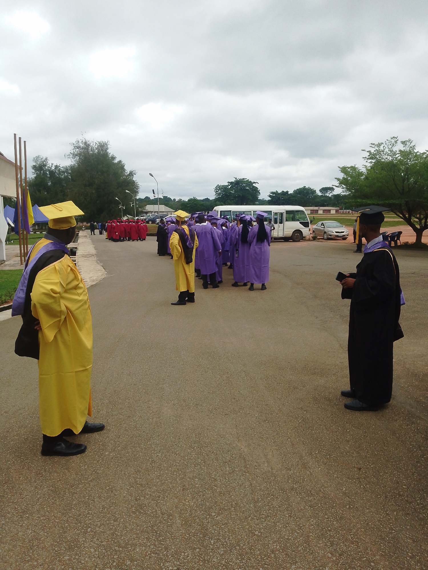 The image depicts an outdoor scene of a graduation ceremony or procession. Several individuals are dressed in academic regalia, including gowns and caps. In the foreground, two individuals are facing away from the camera, one in a yellow gown and cap, and another in a black gown and cap. Behind them, a group of people in purple gowns and caps are gathered, and further in the background, a line of people in red gowns and caps are visible. A white bus and a silver car are parked in the middle ground. The environment appears to be a paved area with trees and a cloudy sky overhead. The overall impression is of a formal academic event taking place outdoors.