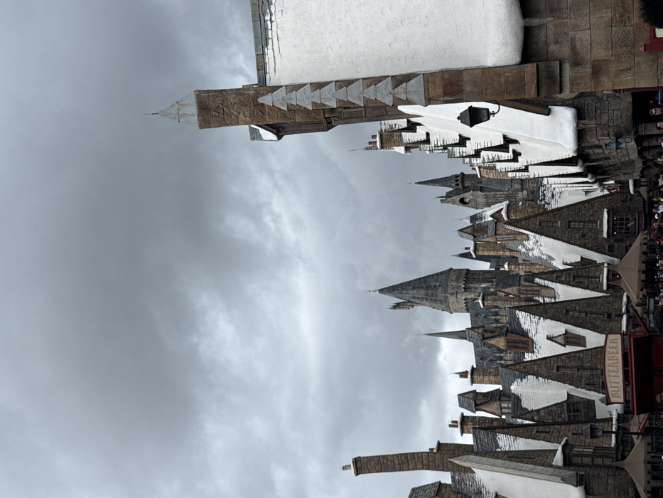 The image displays a detailed view of a themed area within Universal's Islands of Adventure theme park, located in Orlando, Florida, United States of America. The scene depicts "The Wizarding World of Harry Potter - Hogsmeade."
The environment is characterized by an entirely overcast sky, filled with dense, uniform grey clouds, suggesting a dull or potentially rainy day. The predominant subjects are numerous buildings designed in a medieval fantasy architectural style. These structures feature steeply pitched, dark grey roofs, many of which are covered with simulated white snow, creating a winter aesthetic. Stone-textured walls and numerous chimneys of varying heights and designs are also prominent.
In the midground, a street of these buildings is visible, including a red vendor cart or booth prominently displaying a white sign that reads "BUTTERBEER." A large, imposing castle structure, identifiable as Hogwarts Castle, dominates the background with multiple grey spires and towers. To the far right, a tall, narrow, stone-textured building segment rises into the frame, adorned with simulated snow on its roofline and a dark, ornate street lamp affixed to its wall.
At the bottom of the frame, a crowd of indistinct individuals, comprising various people, is visible. They appear to be walking or congregating along a pathway, suggesting active visitor presence in this commercial and recreational area. Dark-colored awnings or canopies extend over sections where people are gathered. No specific interactions between individuals are discernible, and no distinct actions are performed by any single person.