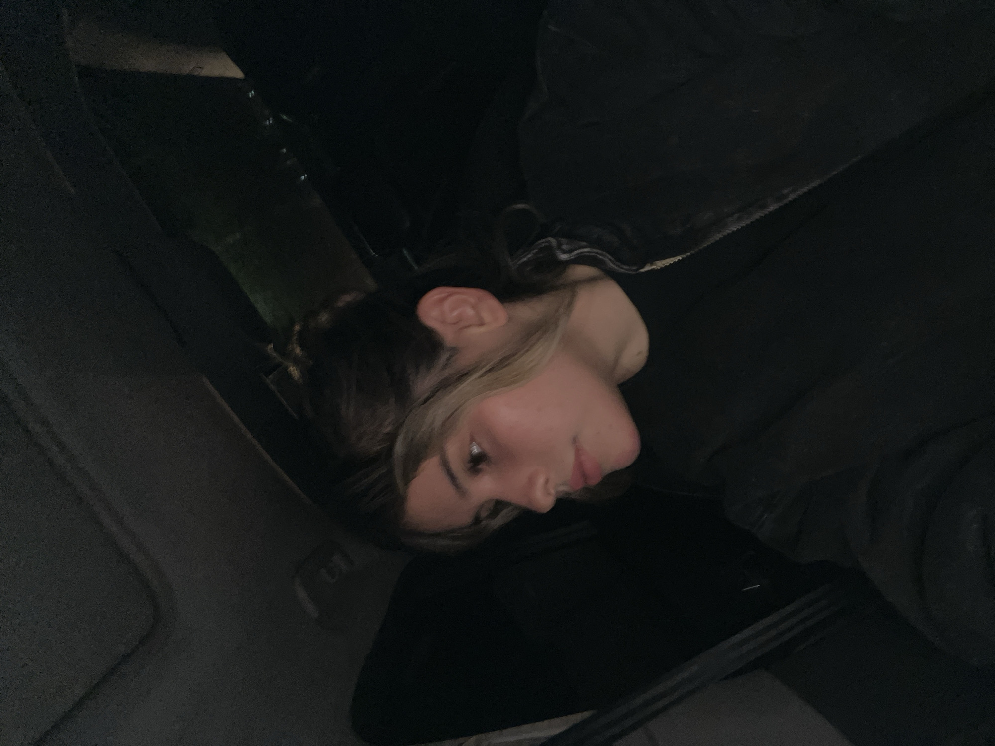 A young woman with blonde highlights in her dark brown hair is seated inside a vehicle, looking towards her left. She is wearing a black jacket over a black shirt. The interior of the vehicle is dark, with the ceiling visible at the top and black upholstery or windows on the sides. The scene suggests a nighttime setting within the city of San Fernando del Valle de Catamarca, Argentina, possibly on a street or highway, given the faint external lights visible in the background.