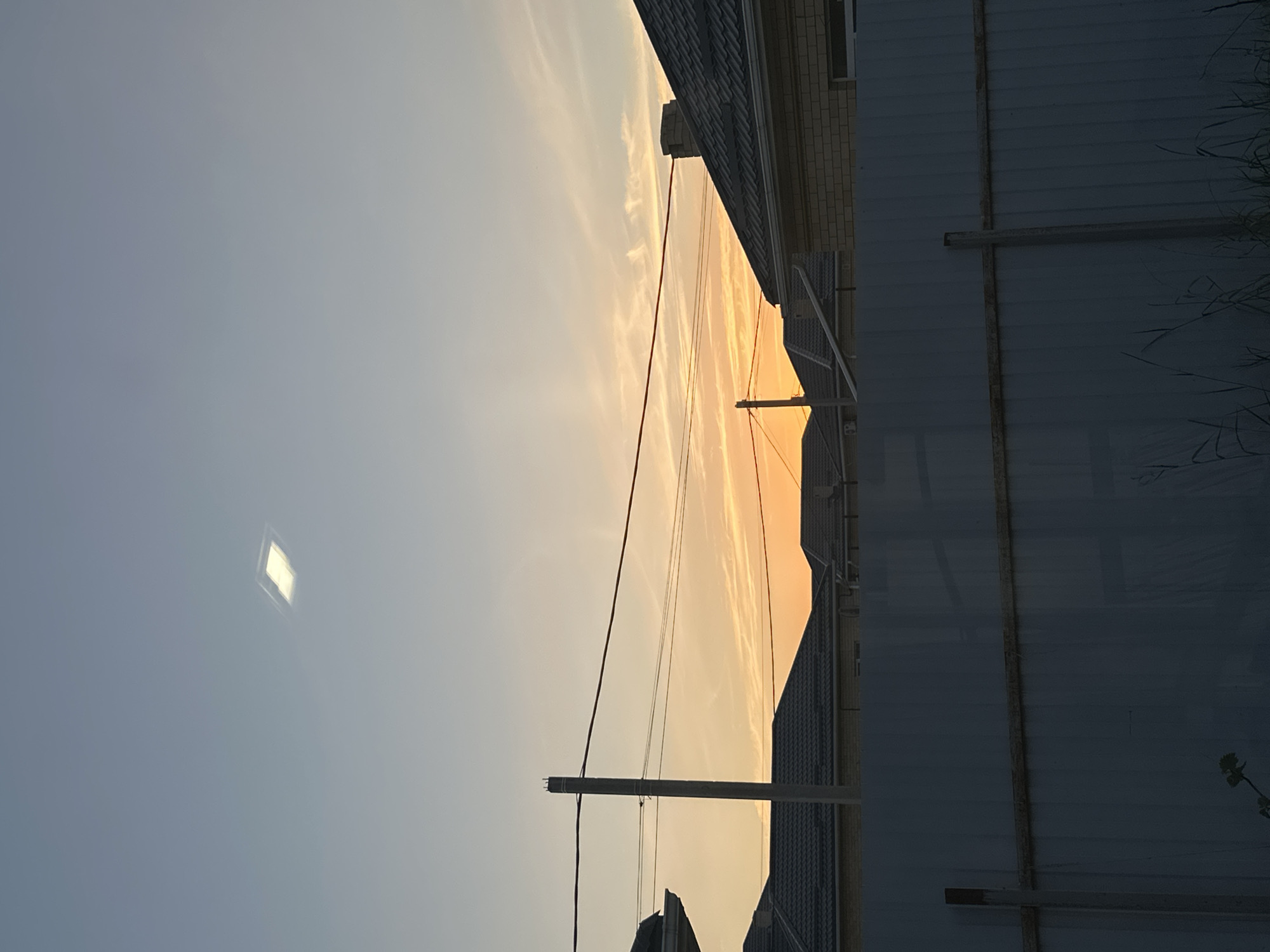 The image depicts a sky scene at dusk, with the sun low on the horizon and casting a warm, orange glow through thin clouds. The sky transitions from a pale blue at the top left to a vibrant orange near the horizon. A solitary, small, bright square object, possibly a light reflection or a distant object, is visible in the upper left portion of the sky.
In the foreground, silhouetted against the sunset, are the rooftops of buildings and a section of a corrugated metal fence. Electrical wires are strung between poles, crossing the frame horizontally. The overall impression is one of a quiet, possibly residential, outdoor setting as daylight fades. The scene evokes a sense of calm and the transition from day to night. There is no visible text in the image.