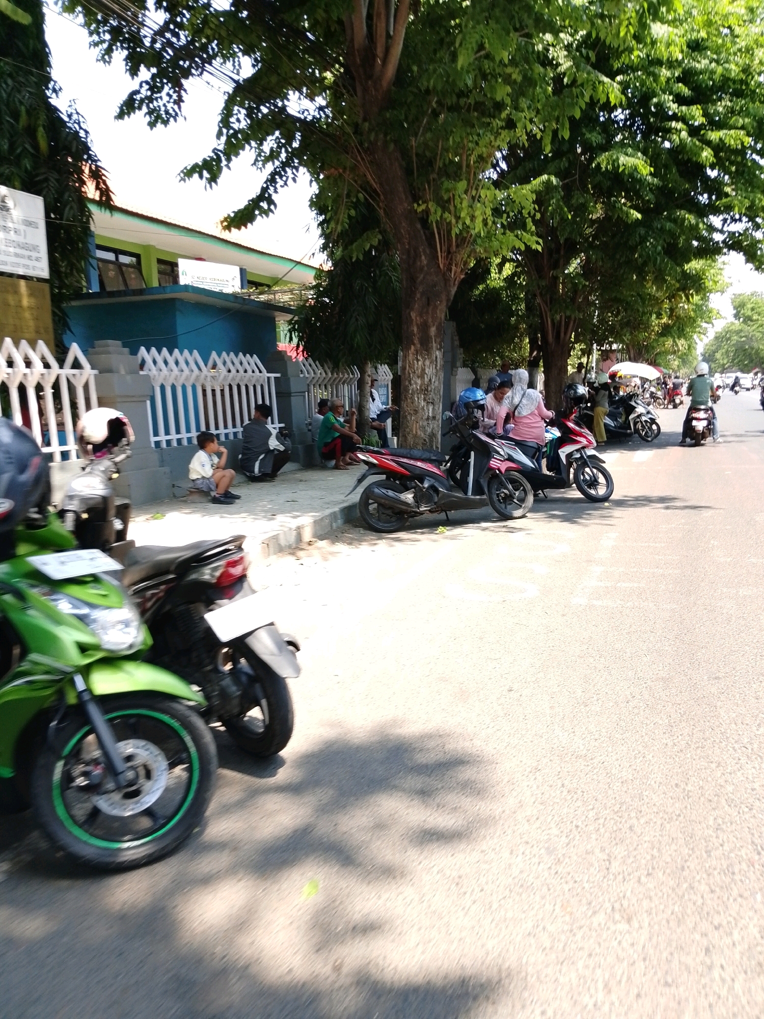 In the city of Pasuruan, Indonesia, a street scene unfolds in front of a building with a blue lower section and white decorative fencing. Several motorcycles are parked on the side of the road, with a bright green motorcycle prominently featured in the foreground. People are seen gathered on the sidewalk, some sitting, others standing, and a few on motorcycles. The environment is sunny with shadows cast by trees on the pavement. One sign visible on the building reads "SD NEGERI KEDUNGSOKO". The overall impression is a typical day in a residential or educational area with local transportation.