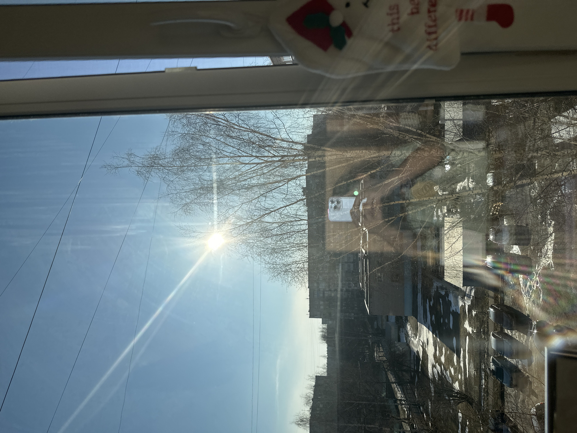 The image is an interior shot taken through a window, with reflections obscuring part of the outside view. The sky is blue with a bright sun. Bare trees are visible against the sky and cityscape. Buildings and ground with patches of snow are present. The location is Izhevsk, Russia. A small Christmas decoration hangs at the top of the window. A reflection of a person holding a phone is also visible.