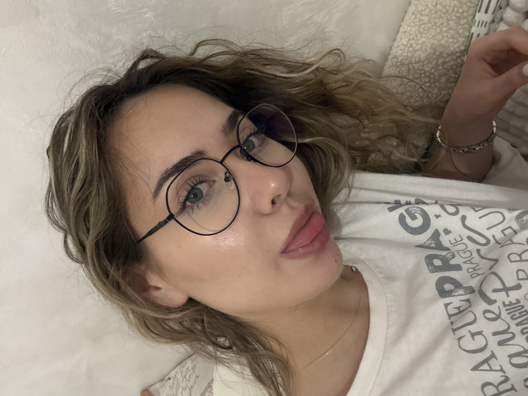 A close-up, high-angle portrait captures a young woman, likely in her late teens or early twenties, reclining indoors, possibly in Blagoevgrad, Bulgaria. She has light green or blue eyes framed by round, black-rimmed glasses, and light brown, wavy hair with subtle blonde highlights. Her eyebrows are well-groomed, and her skin appears clear. Her lips are slightly parted in a pout, with her tongue subtly visible, conveying a playful or alluring expression.
She is wearing a thin, golden chain necklace and a silver-colored link bracelet on her right wrist, which is partially visible. Her attire includes a white t-shirt featuring large, grey, somewhat distressed text repeating the word "PRAGUE" multiple times as part of its design. The background consists of soft, light-colored textures, resembling a pillow or blanket, suggesting a relaxed, indoor setting like a bedroom or living room. The overall lighting is soft, indicating it could be daytime indoors or under artificial light. No specific weather conditions are discernible.