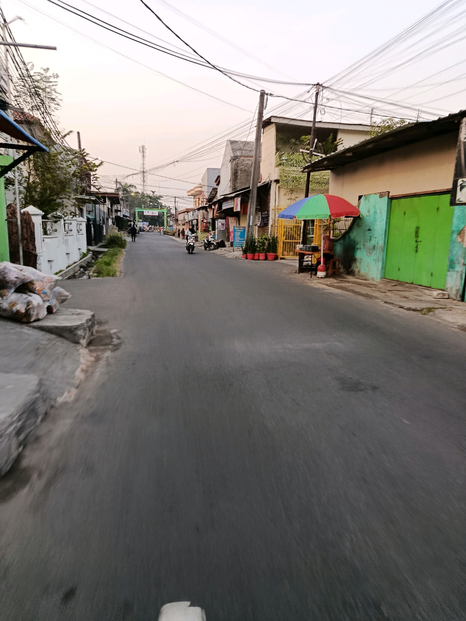 The image shows a street scene in the city of Bekasi, Indonesia, at what appears to be dawn or dusk. The street is paved and appears to be a local road, with buildings lining both sides. In the foreground, a motorcycle is visible on the right side of the road, with a rider wearing a headscarf. Further down the street, another motorcycle is seen, and there are a few pedestrians.
On the left side of the street, there are white walls with some trees and vegetation. Bags of what appear to be garbage or materials are piled on the side of the road in the lower left corner. On the right side of the street, a vendor stall with a colorful umbrella is set up, and a person is seated behind it. The buildings on this side of the street appear to be shops or homes.
Overhead, a network of power lines and cables crisscrosses the sky, typical of many urban areas in Indonesia. In the distance, a cell tower is visible. The overall atmosphere of the image is that of a typical, lived-in urban environment with a mixture of residential and commercial activity.