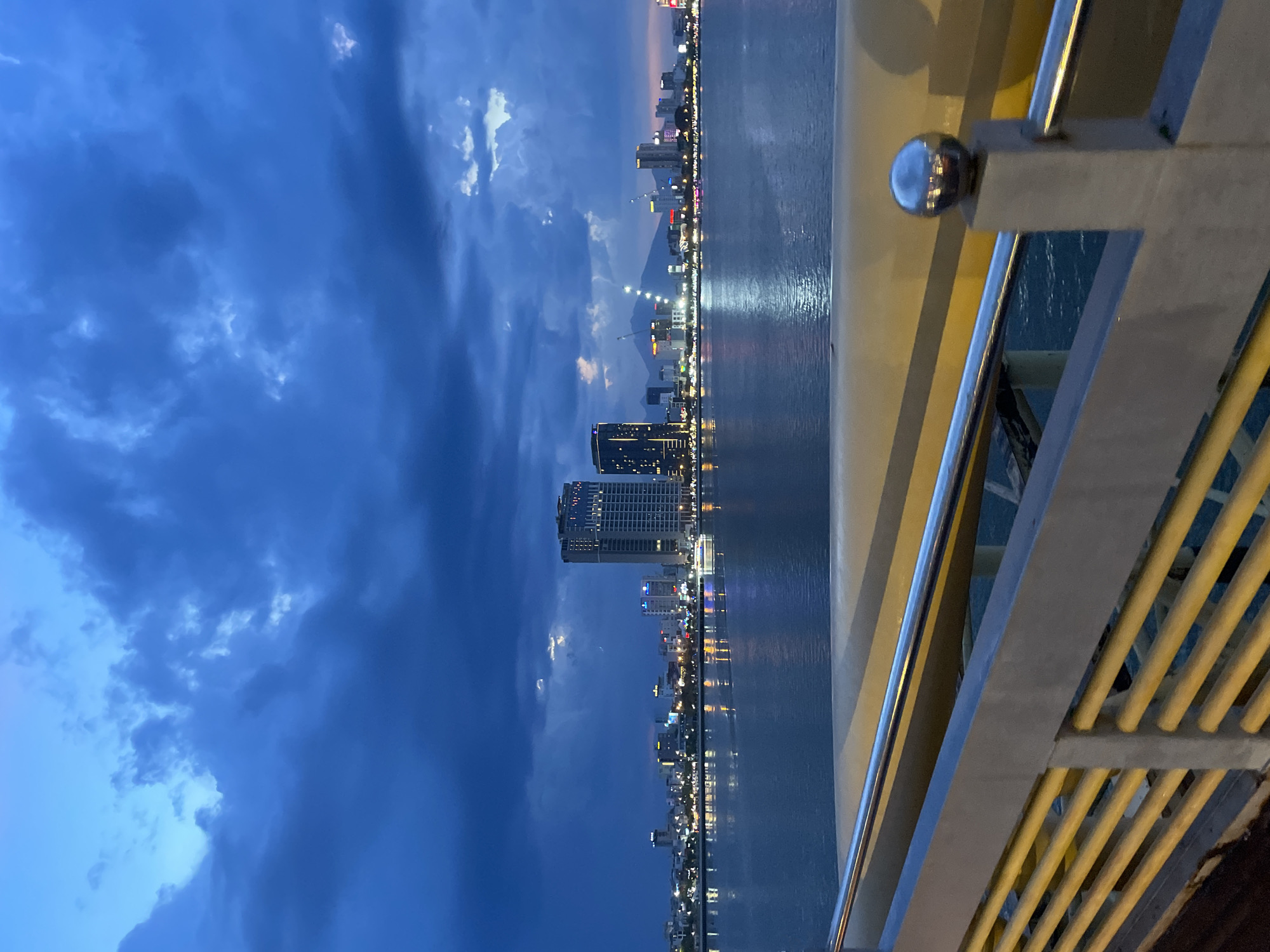 The image depicts a twilight view of the Da Nang cityscape and coastline. The scene is dominated by a vast expanse of deep blue sky with scattered, dramatic clouds, suggesting a recent or impending weather change. A shimmering body of water, likely the sea, occupies the lower portion of the frame, reflecting the lights of the city.
The cityscape is characterized by numerous buildings of varying heights, illuminated with a mix of colorful lights, indicating a bustling urban environment. Prominent among these are several tall, modern high-rise buildings, some featuring illuminated facades and windows. The reflection of these lights on the water creates a mesmerizing visual effect. In the distance, a faint mountain range is visible, adding depth to the landscape.
The foreground is partially occupied by a yellow and gray structure, possibly a railing or part of a bridge, with metallic elements and yellow, ribbed bars. A spherical, metallic object is attached to the railing structure, reflecting the ambient light. The perspective suggests the viewer is positioned on an elevated vantage point, looking out over the city and sea.
The time of day is clearly evening or dusk, as indicated by the deep blue sky and the artificial lights of the city becoming prominent. The weather appears dynamic, with the clouds hinting at a dramatic sky. While no people are explicitly visible, the illuminated city suggests ongoing activity. The overall mood is serene yet vibrant, capturing the dynamic beauty of a coastal city at twilight. No visible text is present in the image.