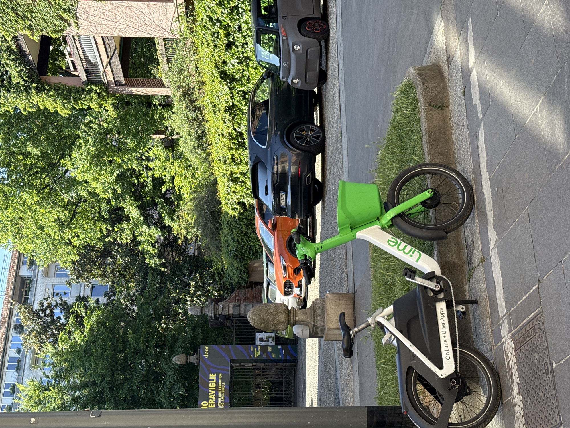 The image shows a Lime scooter parked on the sidewalk in Milan, Italy. A row of cars is parked along the curb, including a gray Citroën Ami, a black Porsche Cayenne with license plate "CD414MM", and an orange car. Behind the cars are buildings, trees, and climbing vines. A sign visible partially from behind a stone fence advertises an exhibition called "10 EraViglie." The lighting suggests it's daytime. The street surface is made of square concrete or stone blocks.