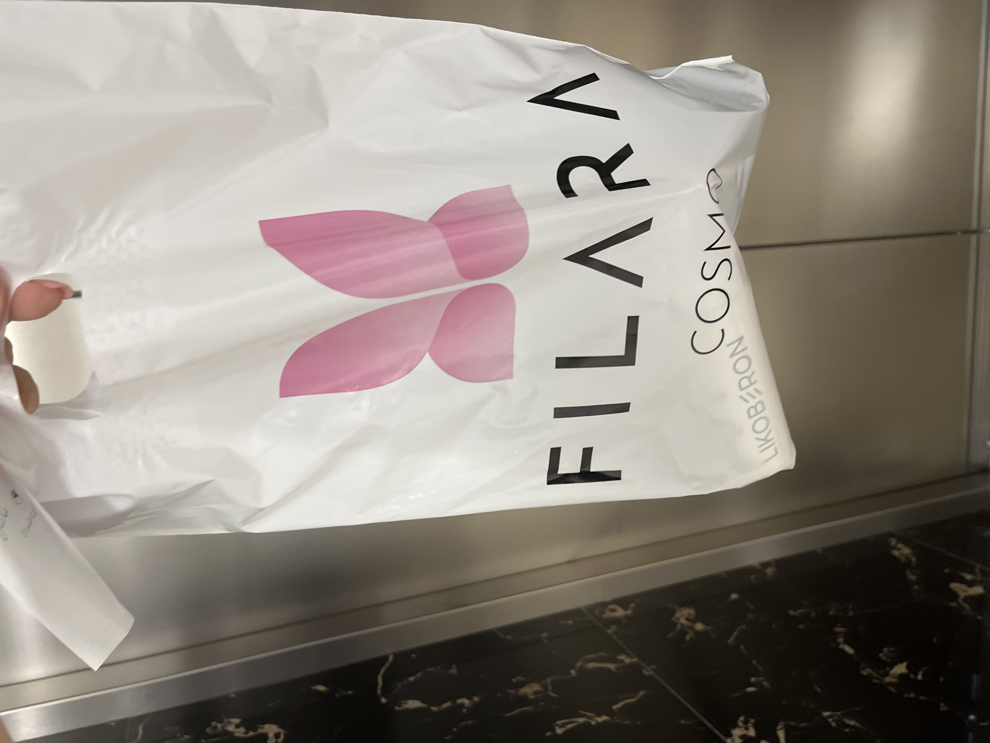 A person is holding a white plastic shopping bag with a pink butterfly logo and black text. The bag is branded "FILARA" and also displays the words "LIKOBERON COSMO". The background appears to be an elevator interior, with a dark patterned wall on the left and a brushed metal surface on the right. The lighting suggests an indoor setting.