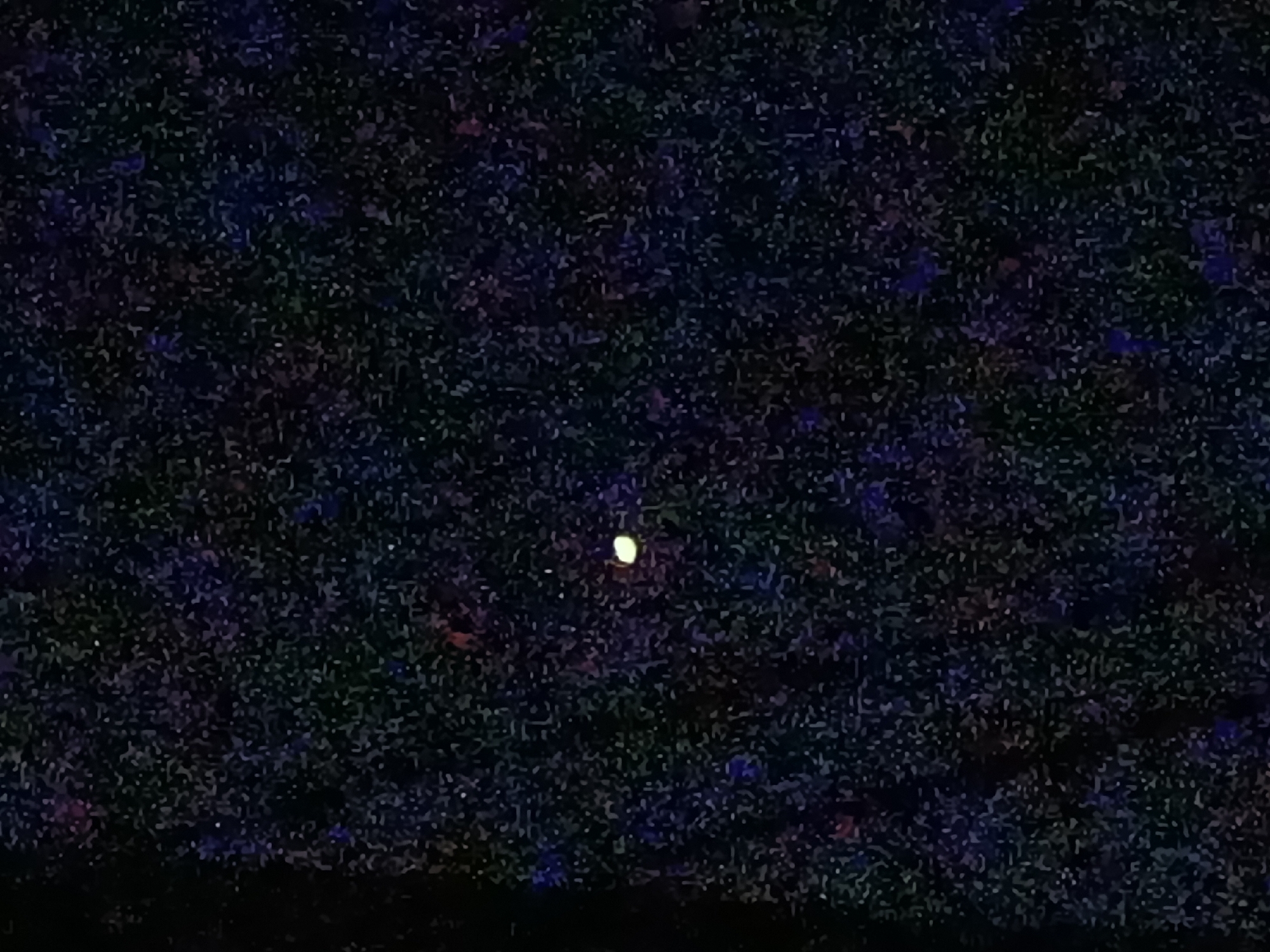 The image shows a dark night sky filled with numerous small, indistinct specks of light, resembling stars. In the center of the frame, a single, slightly larger, circular object glows with a yellowish-white light, appearing to be a celestial body. The sky has a mottled appearance with hints of dark blues, purples, and greens, suggesting a distant or perhaps slightly hazy atmosphere. The bottom edge of the image is darker, indicating the presence of a foreground element, likely the silhouette of the earth's surface, possibly a landscape or city skyline. The scene is devoid of any clear human figures or specific man-made structures that would allow for precise identification of the location within Kyzyl-Suu, Kyrgyzstan. The overall impression is that of an astronomical observation or a nighttime landscape.