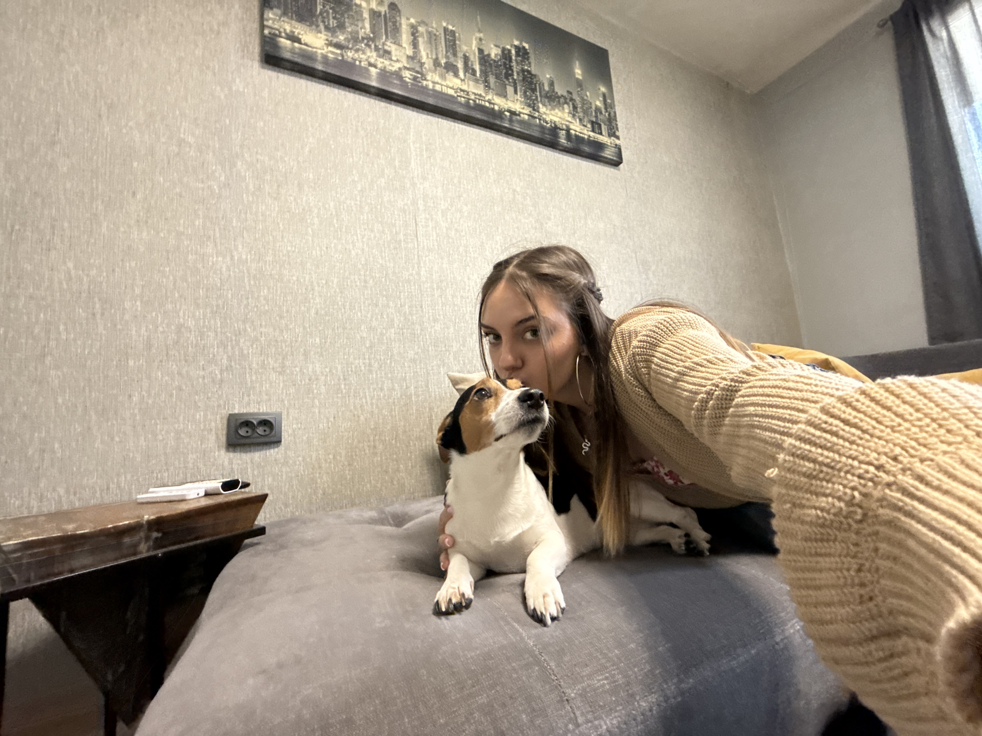 A woman is taking a selfie with a dog. The dog, a Jack Russell Terrier, sits on a grey upholstered surface. The woman is wearing a beige sweater and silver hoop earrings, and she appears to be kissing the dog on the head. In the background, there's a cityscape picture hanging on a grey wall, a dark-colored table with a white remote, and a dark-colored curtain partially visible. The location is likely indoors in a residential setting. There is no information to identify the location as Sofia, Bulgaria.